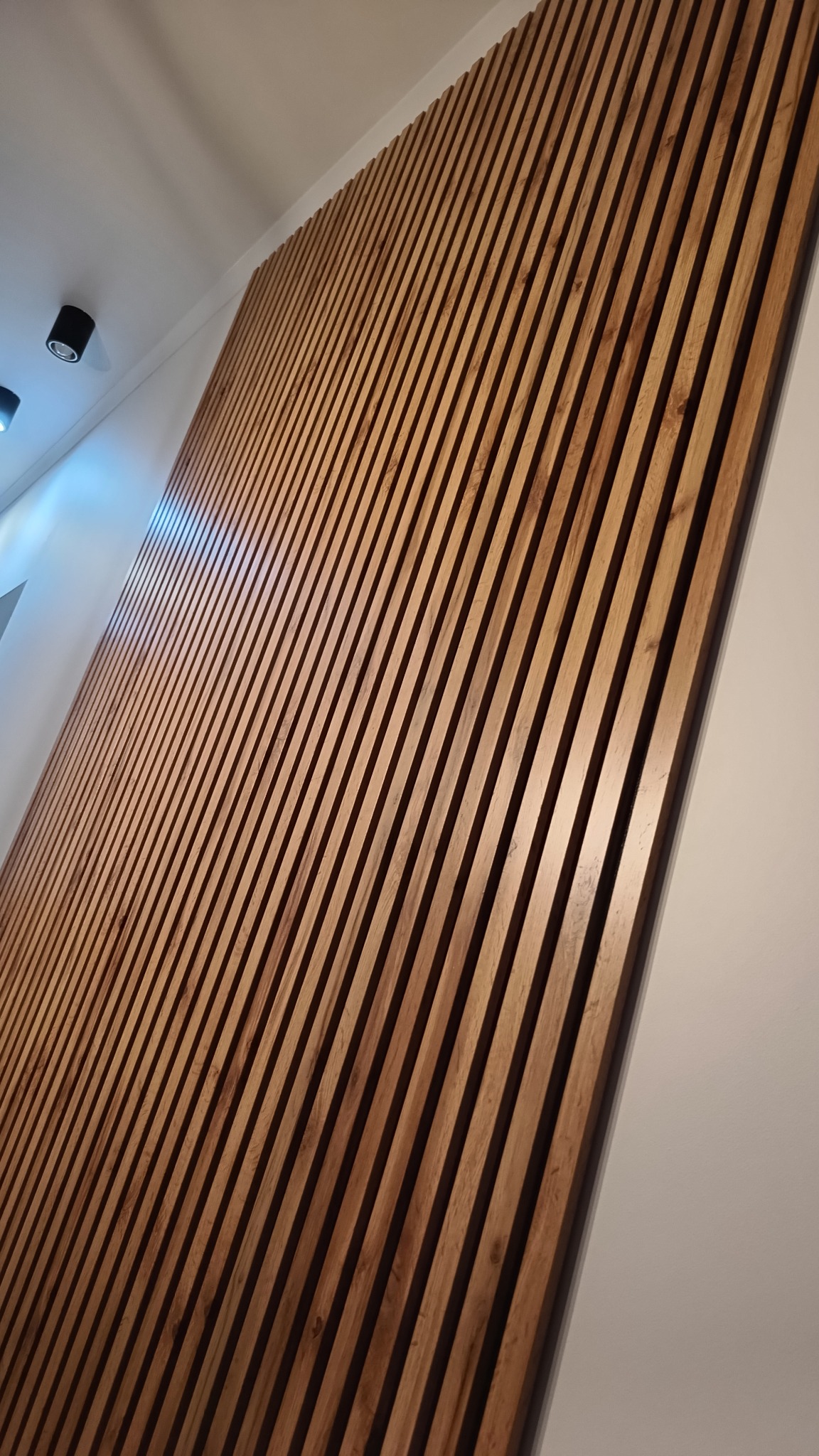 Dekoracyjna ściana z drewnianych listew w ciepłym odcieniu brązu, zamontowana skośnie na białej ścianie, oświetlona przez punktowe lampy sufitowe.