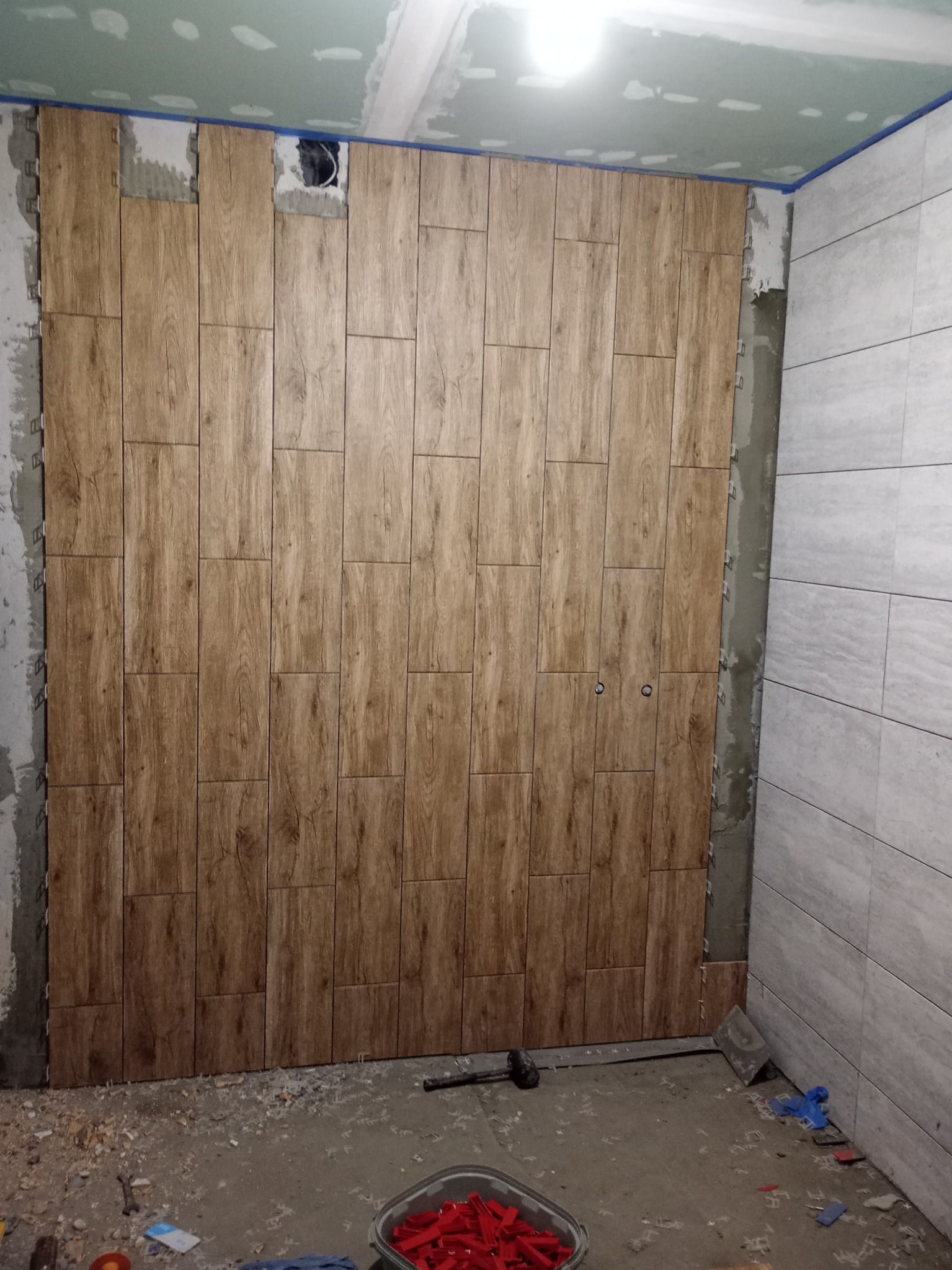 Ściana łazienki w trakcie układania płytek imitujących drewno, widoczne krzyżyki dystansowe i narzędzia glazurnicze. Częściowo wykończona ściana z jasnych płytek.