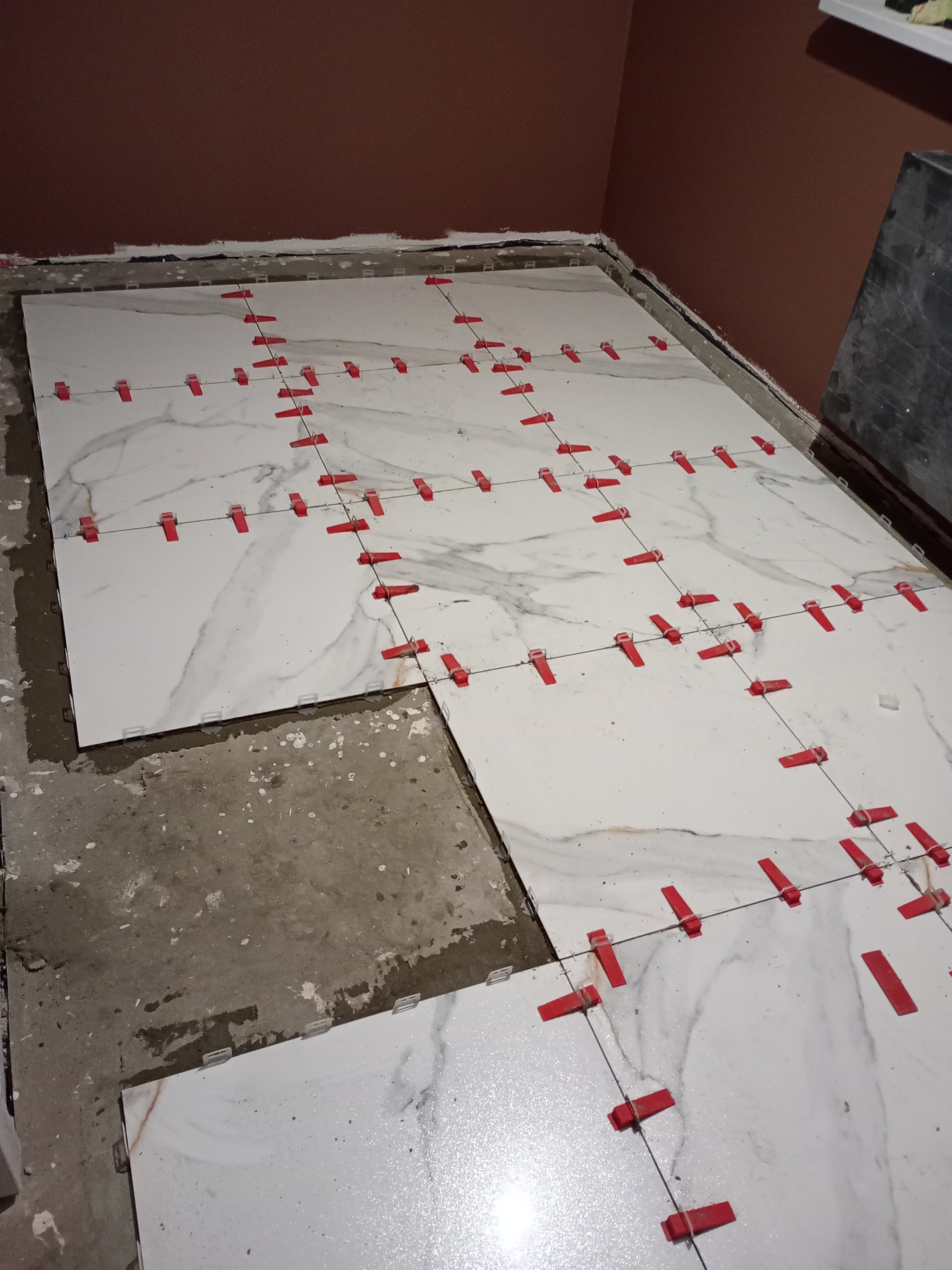 Układanie białych płytek podłogowych z marmurowym wzorem, z czerwonymi klipsami poziomującymi, na nierównej podłodze w pomieszczeniu z brązową ścianą.