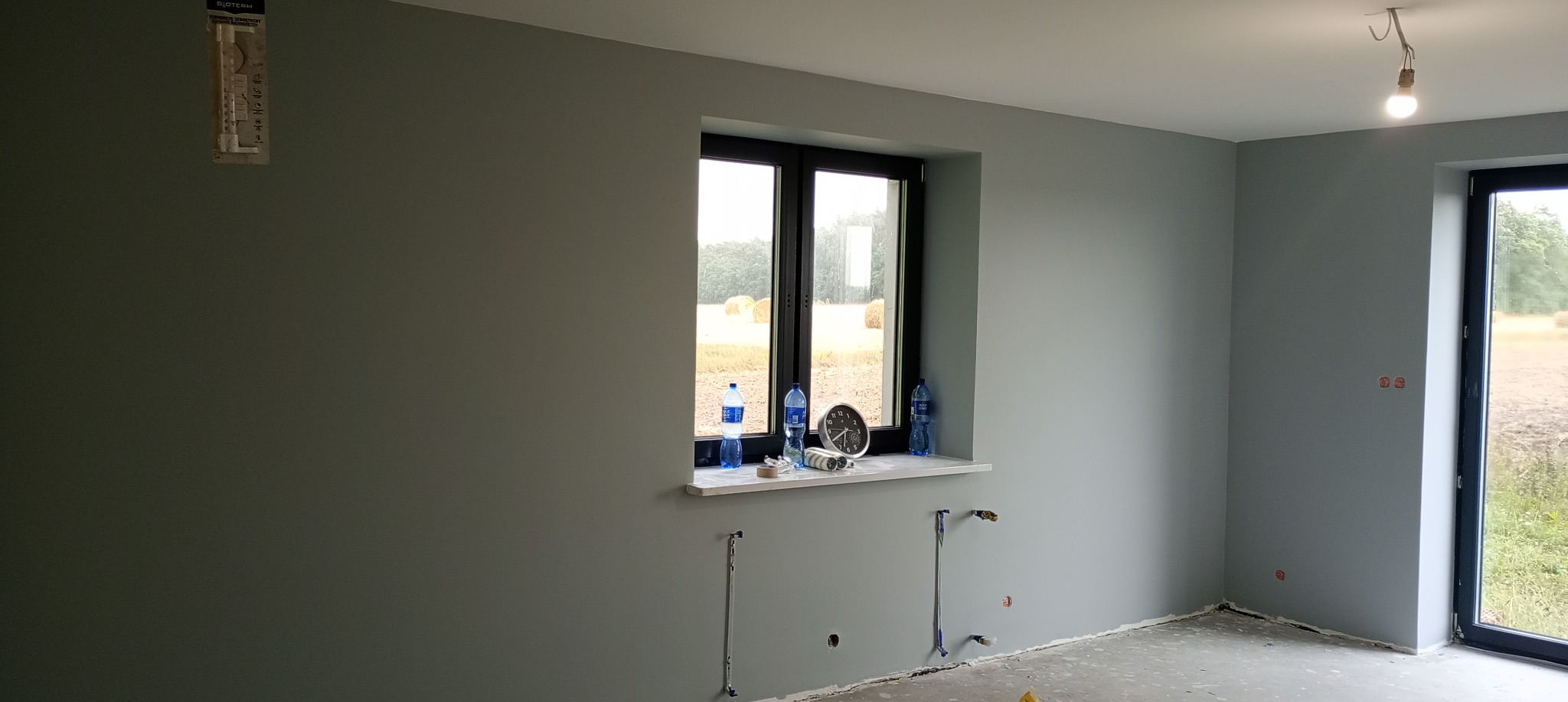 Świeżo pomalowane wnętrze w odcieniach szarości, z widocznymi instalacjami podtynkowymi i oknem z widokiem na pole. Na parapecie butelki wody i zegar.