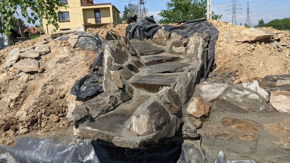 Budowa kaskady wodnej z kamienia łupanego w ogrodzie, widoczne schodki i zabezpieczenie folią. W tle dom i słupy wysokiego napięcia.