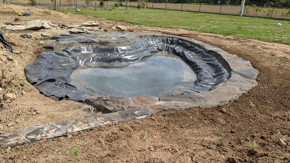 Oczko wodne w trakcie budowy: wykopany dół wyłożony folią, obłożony kamieniami, częściowo wypełniony wodą. W tle trawnik i drzewa.