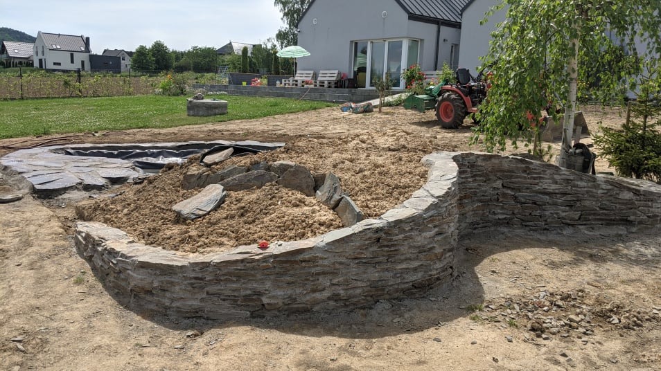 Budowa oczka wodnego z kamienia: murowana obudowa w kształcie półkola, wypełniona piaskiem, w tle traktor i dom. Etap prac w słoneczny dzień.