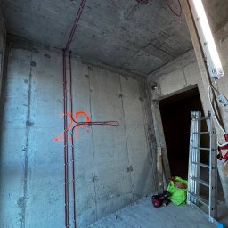 Apem - Surowy betonowy pokój z instalacją elektryczną w czerwonych rurach, przymocowaną metalowymi uchwytami do ściany i sufitu. Widoczna drabina i narzędzia.