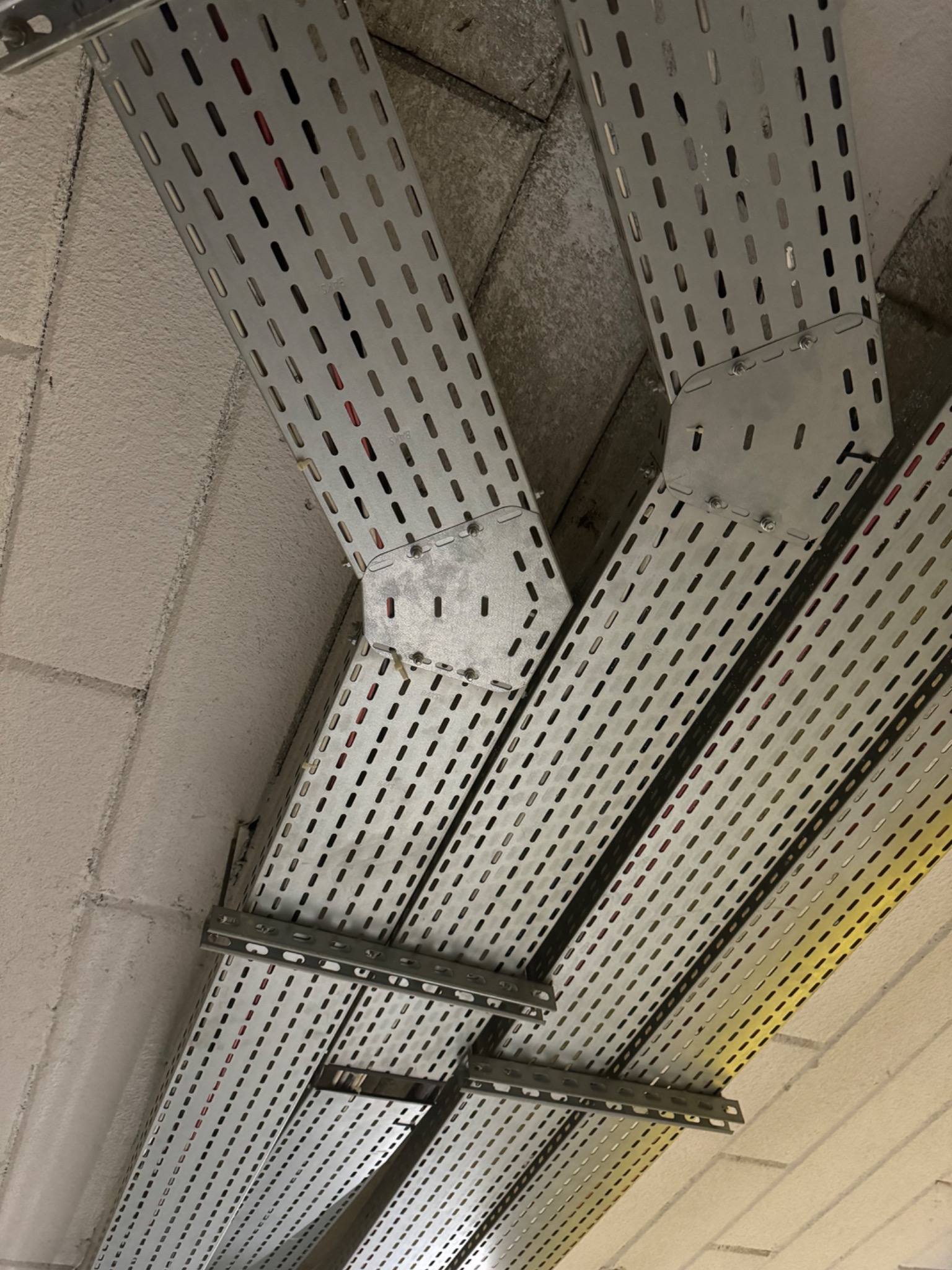 Metalowe koryta kablowe, perforowane, zamontowane pod sufitem z betonu komórkowego. Widoczne łączenia i wsporniki. Kable wewnątrz koryt. Ujęcie z dołu.