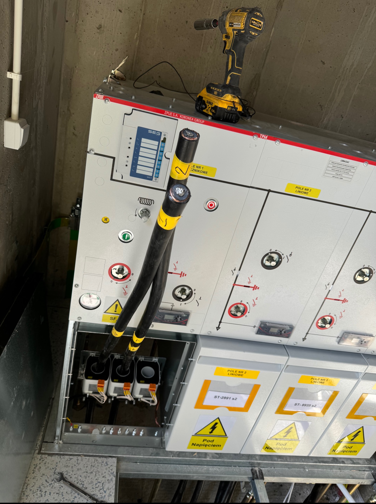 Rozdzielnica elektryczna z podłączonymi kablami, widoczne oznaczenia ostrzegawcze i narzędzie pracy - wiertarka. Instalacja w szarej betonowej wnęce.