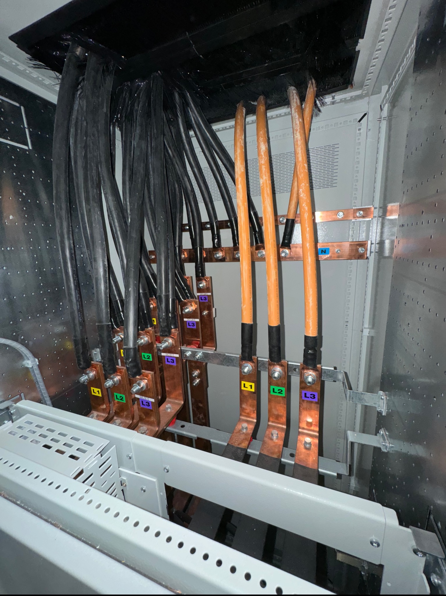 Wnętrze szafy elektrycznej: kable zasilające (czarne) i przewody neutralne (pomarańczowe) podłączone do miedzianych szyn zbiorczych oznaczonych L1, L2, L3 i N.