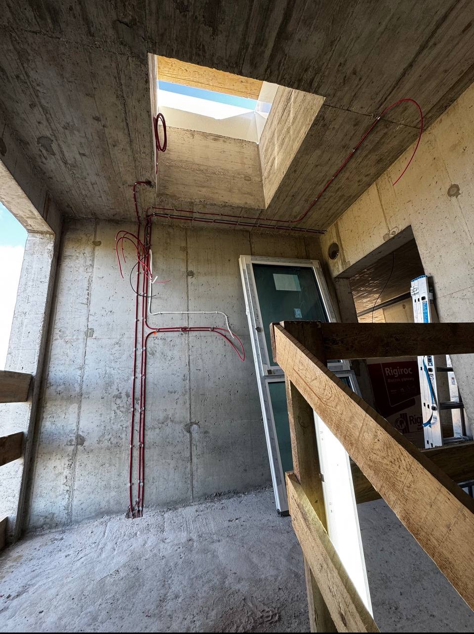 Wewnętrzne ujęcie surowego budynku w trakcie instalacji elektrycznej, z widocznymi czerwonymi przewodami biegnącymi po betonowych ścianach i suficie. W tle okno dachowe.