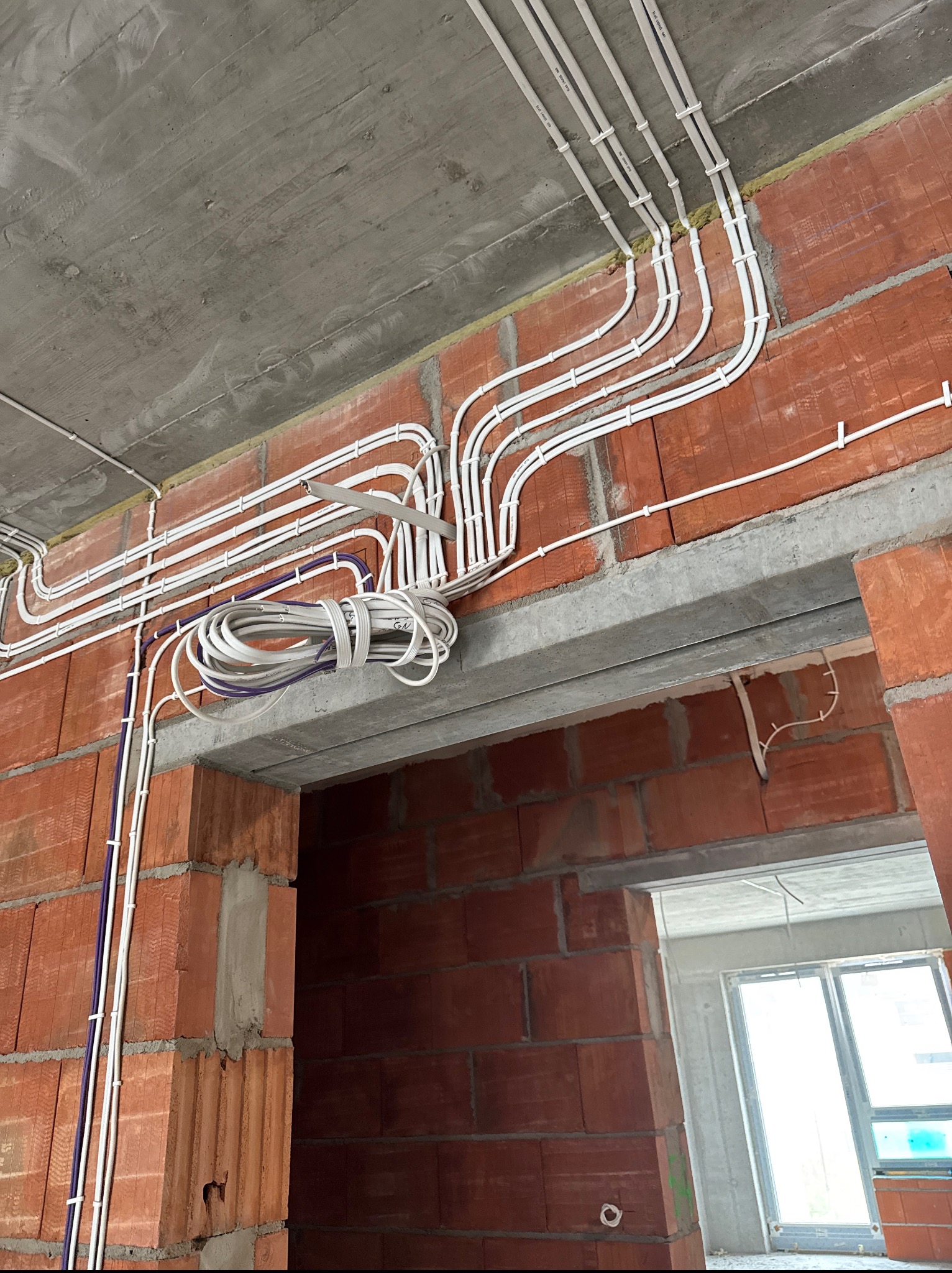 Białe rury instalacji elektrycznej prowadzone po ceglanej ścianie i betonowym suficie w surowym wnętrzu budynku w trakcie budowy.