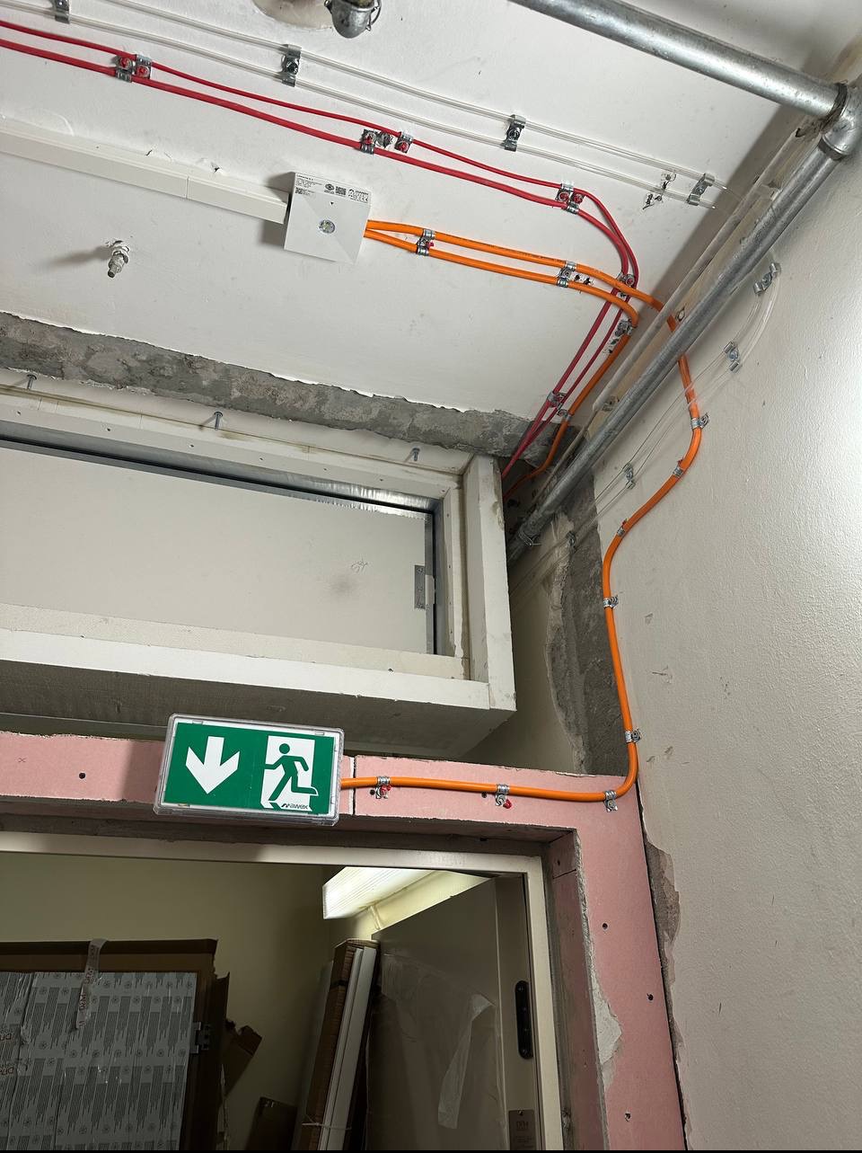 Instalacja elektryczna: rury w kolorach czerwonym i pomarańczowym, przymocowane do sufitu i ściany. Widoczny znak ewakuacyjny nad drzwiami.