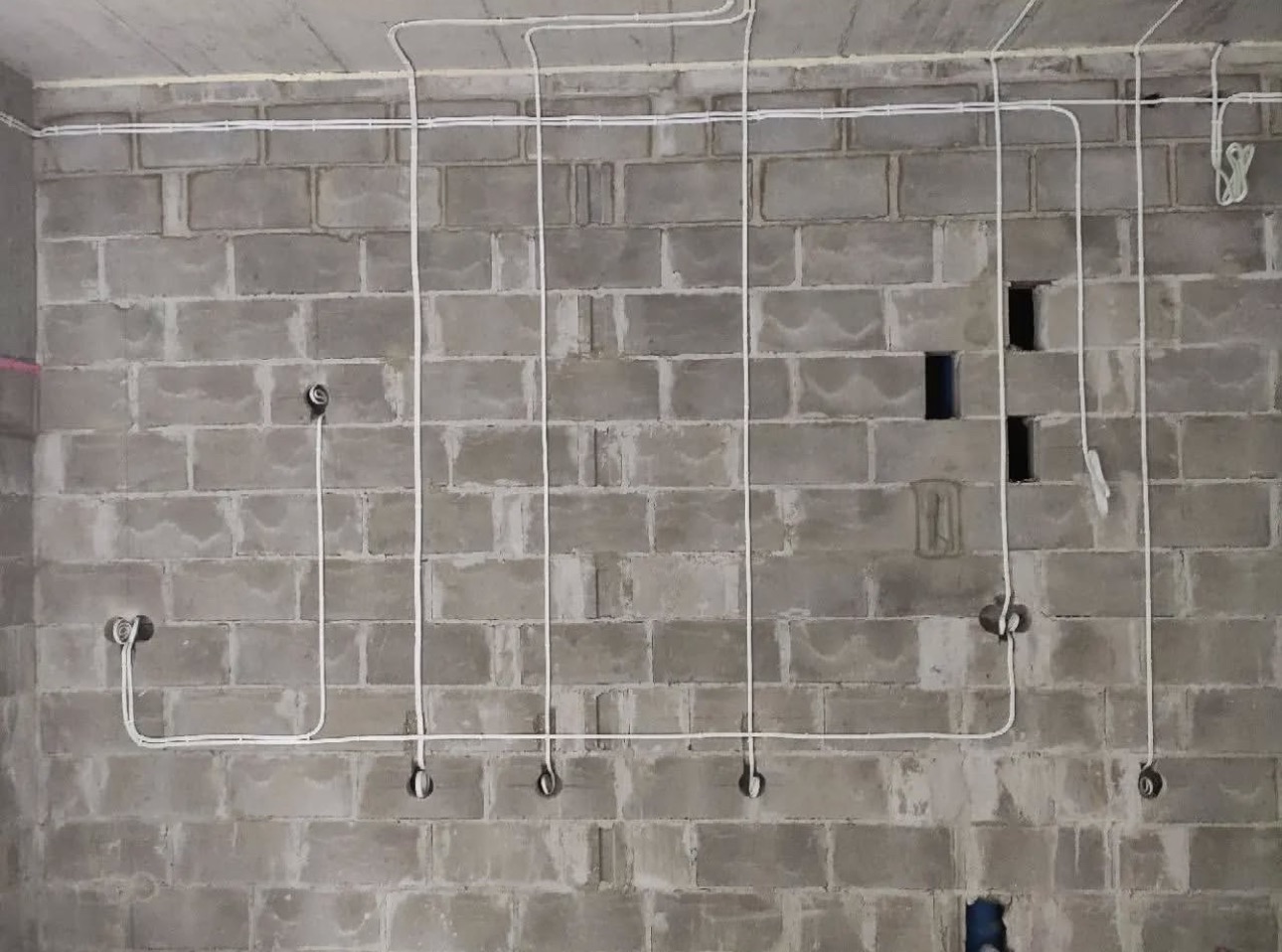 Szara ściana z betonu komórkowego z poprowadzoną białą instalacją elektryczną. Widoczne puszki instalacyjne i przewody ułożone wzdłuż ściany i pod sufitem.