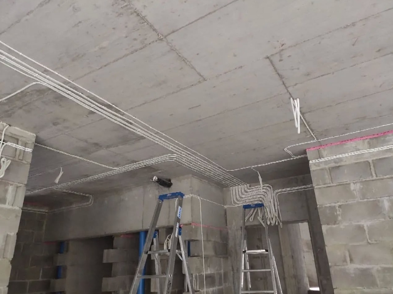 Białe kable elektryczne ułożone pod betonowym sufitem w surowym wnętrzu. Dwie drabiny aluminiowe stoją pod instalacją. Widoczne ściany z bloków.