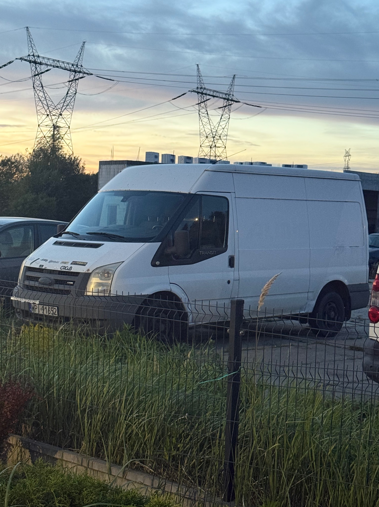 Biały bus Ford Transit zaparkowany przy ogrodzeniu, na tle słupów wysokiego napięcia i zachodzącego słońca. Widoczna tablica rejestracyjna i częściowo inne pojazdy.