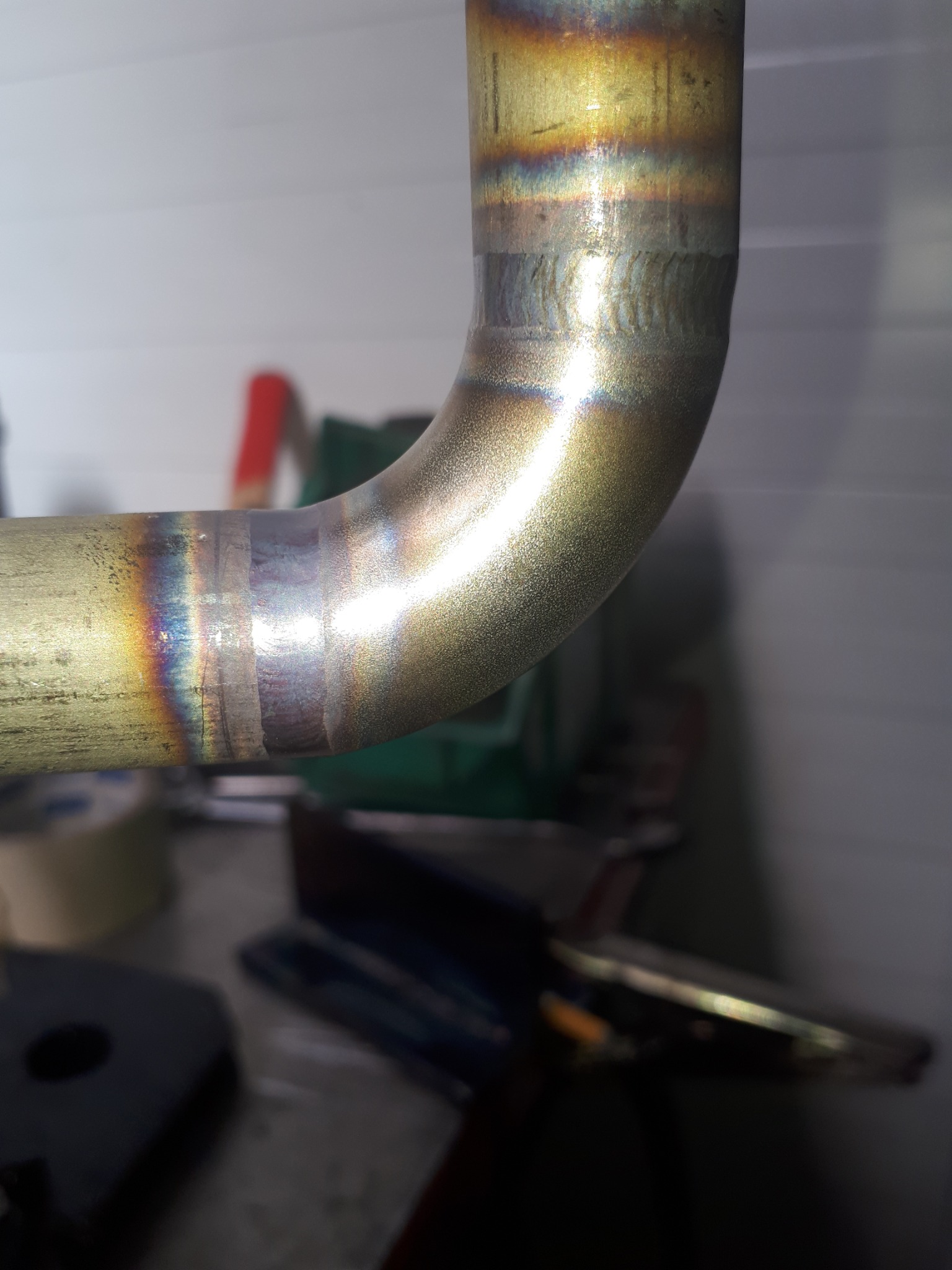 Szczegółowe ujęcie spawu na zagiętej, metalowej rurze. Widoczne ślady ciepła i kolory tęczy na spoinie. W tle warsztatowe otoczenie z narzędziami.