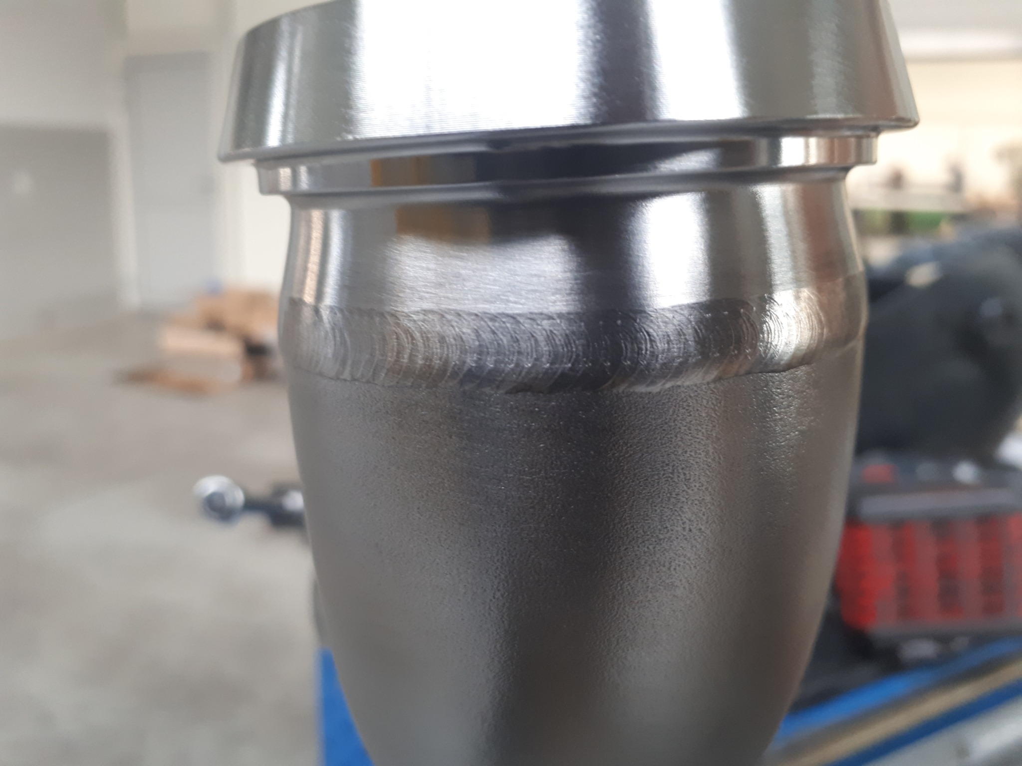 Szlifowany element metalowy po obróbce, widoczne ślady spawania, zarys sprzętu warsztatowego w tle, oświetlenie naturalne, detal.