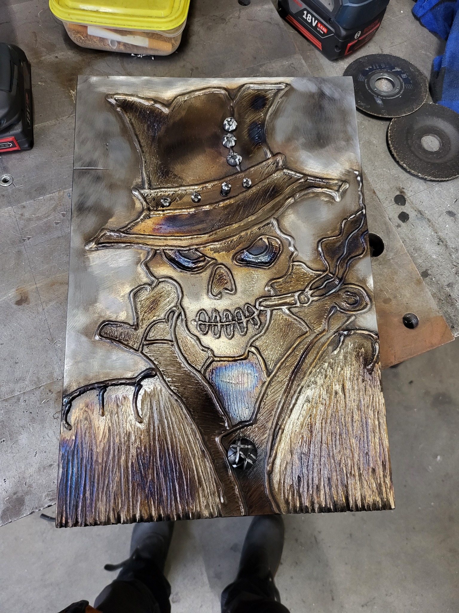 Metalowa płaskorzeźba przedstawiająca czaszkę w kapeluszu z cygarem, wykonana techniką spawania, z widocznymi śladami obróbki i narzędziami w tle.