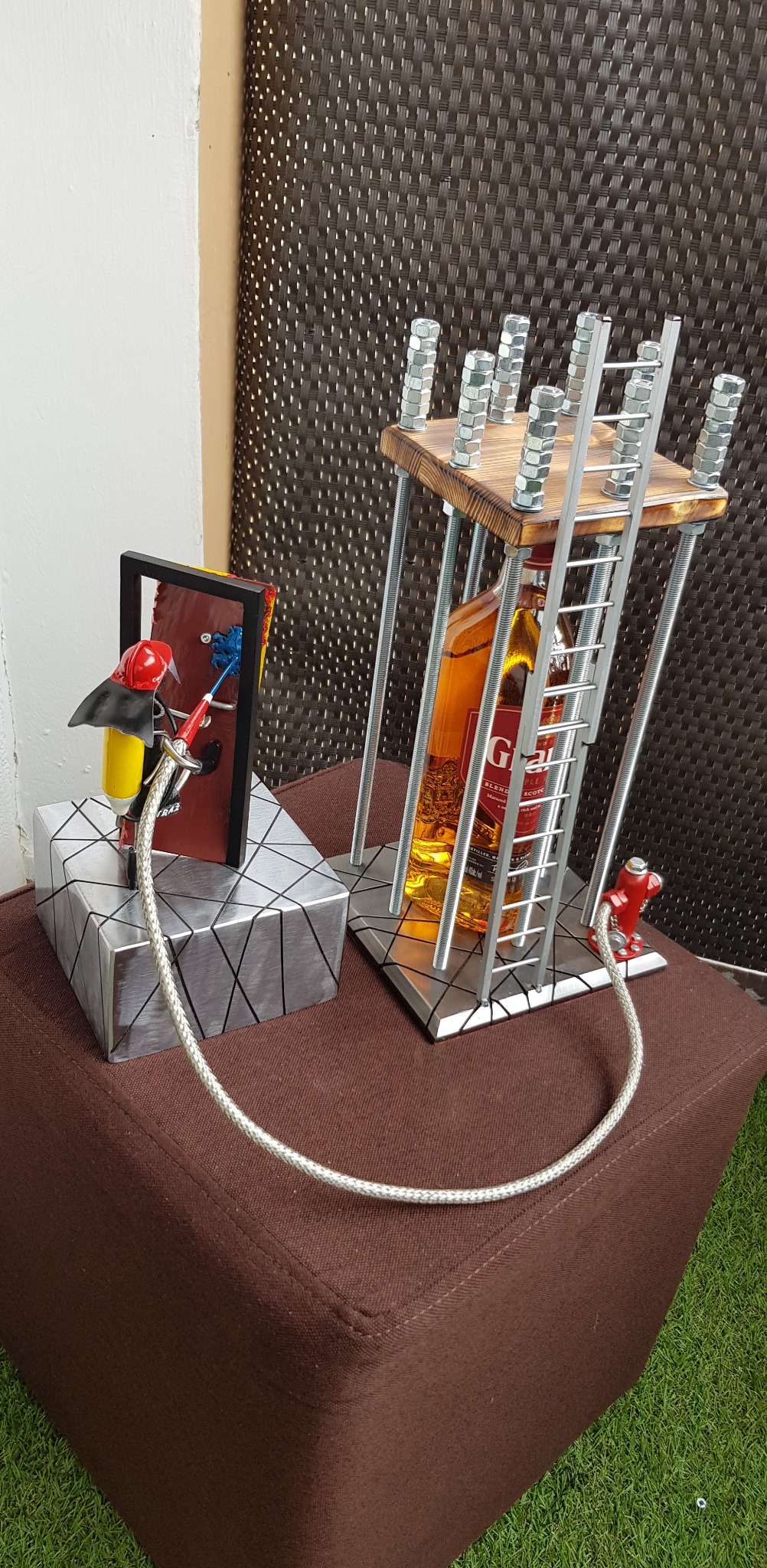 Metaloplastyka: figurka strażaka z wężem i konstrukcja z drabinką otaczająca butelkę, całość na tle brązowej pufy i zielonej trawy.