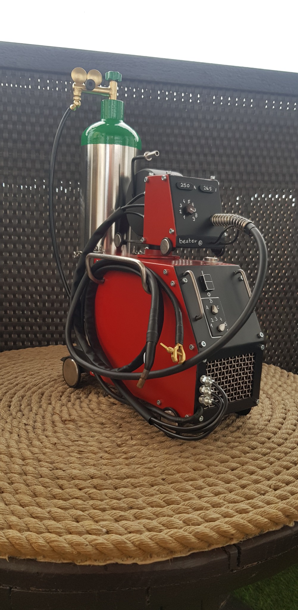 Czerwono-czarna spawarka Bester z butlą gazową na stoliku z liny jutowej, na tle plecionej ściany. Widoczne przewody, pokrętła regulacji i detale konstrukcyjne.