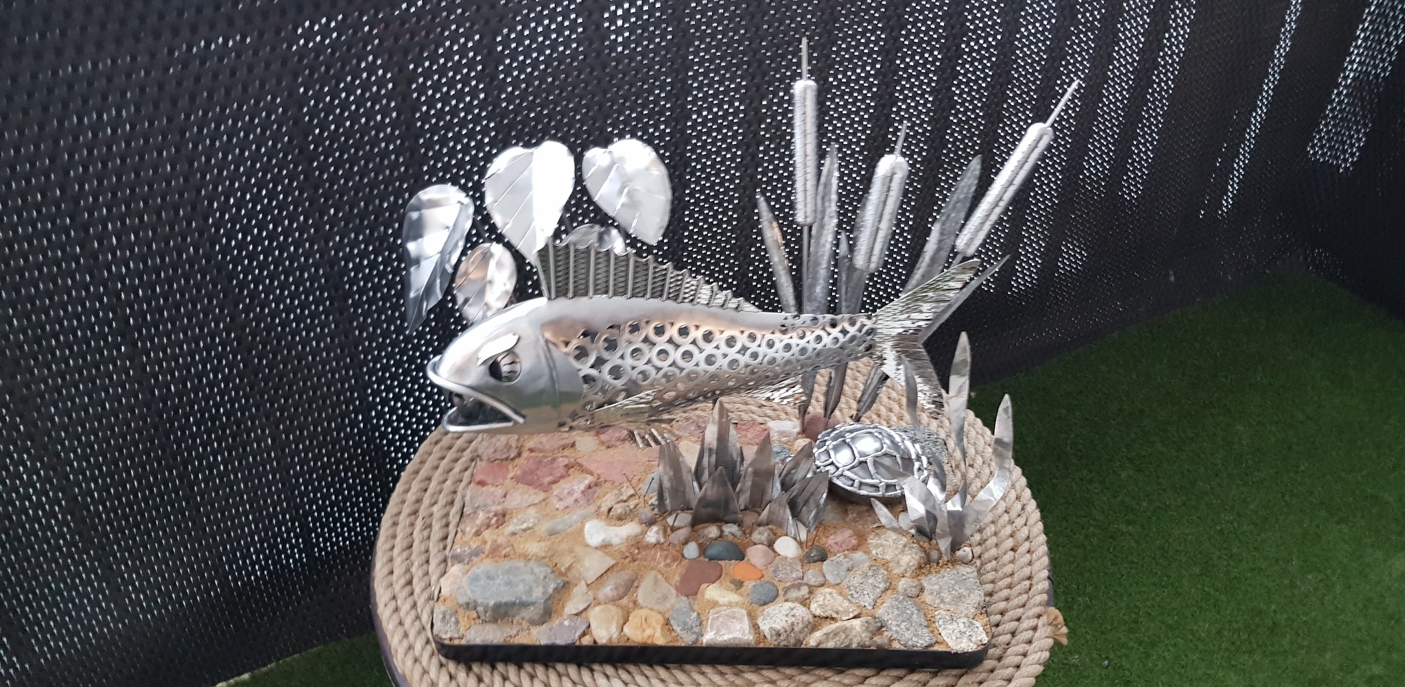 Dekoracyjna rzeźba: metalowa ryba z otworami, trzcina i żółw na podstawie z kamieni, całość na postumencie z liny, na tle czarnej siatki.