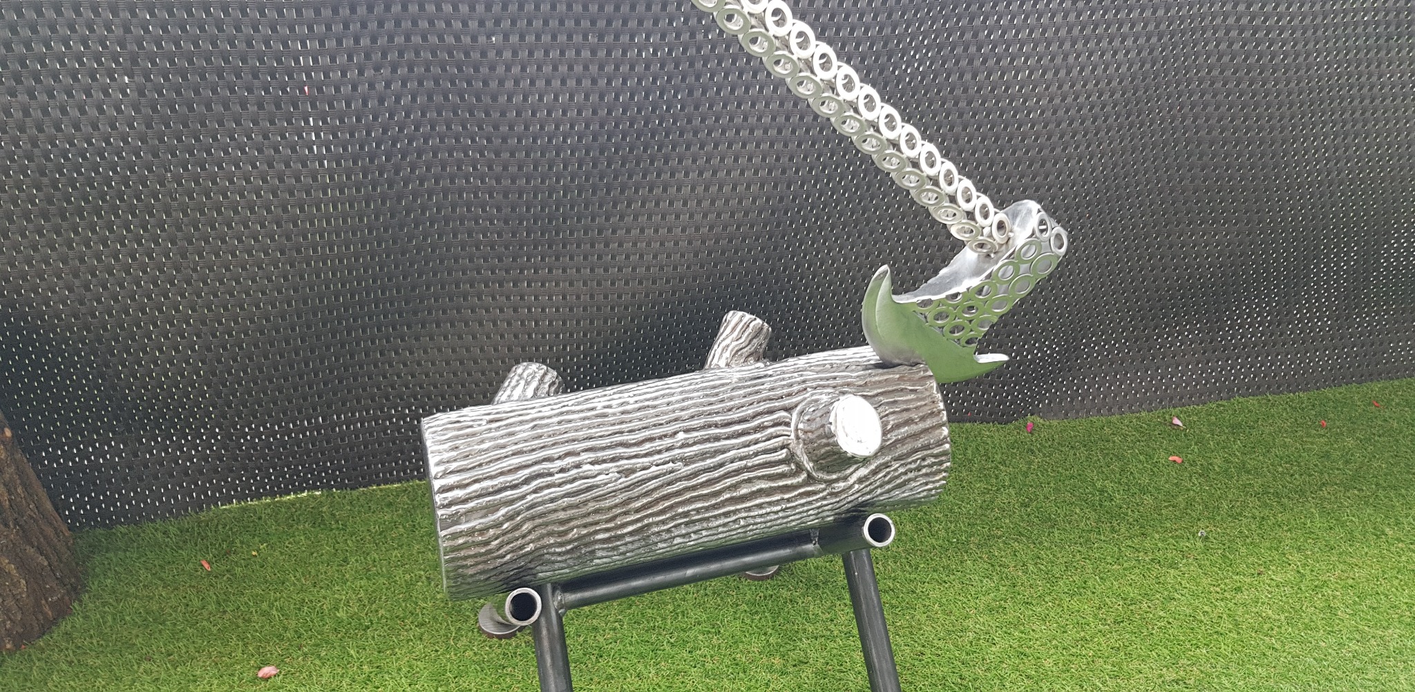 Metalowa rzeźba przedstawiająca pień drzewa z siekierą na tle czarnej maty i sztucznej trawy. Nietypowa forma artystyczna.