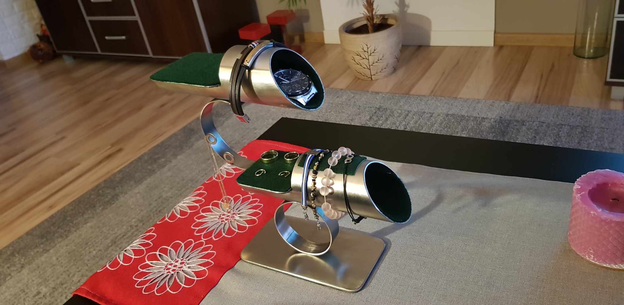 Metalowy stojak na biżuterię z zegarkiem, pierścionkami i bransoletkami na tle wzorzystego obrusa i świecy. Nietypowy design, srebrny kolor, zielone wkładki.