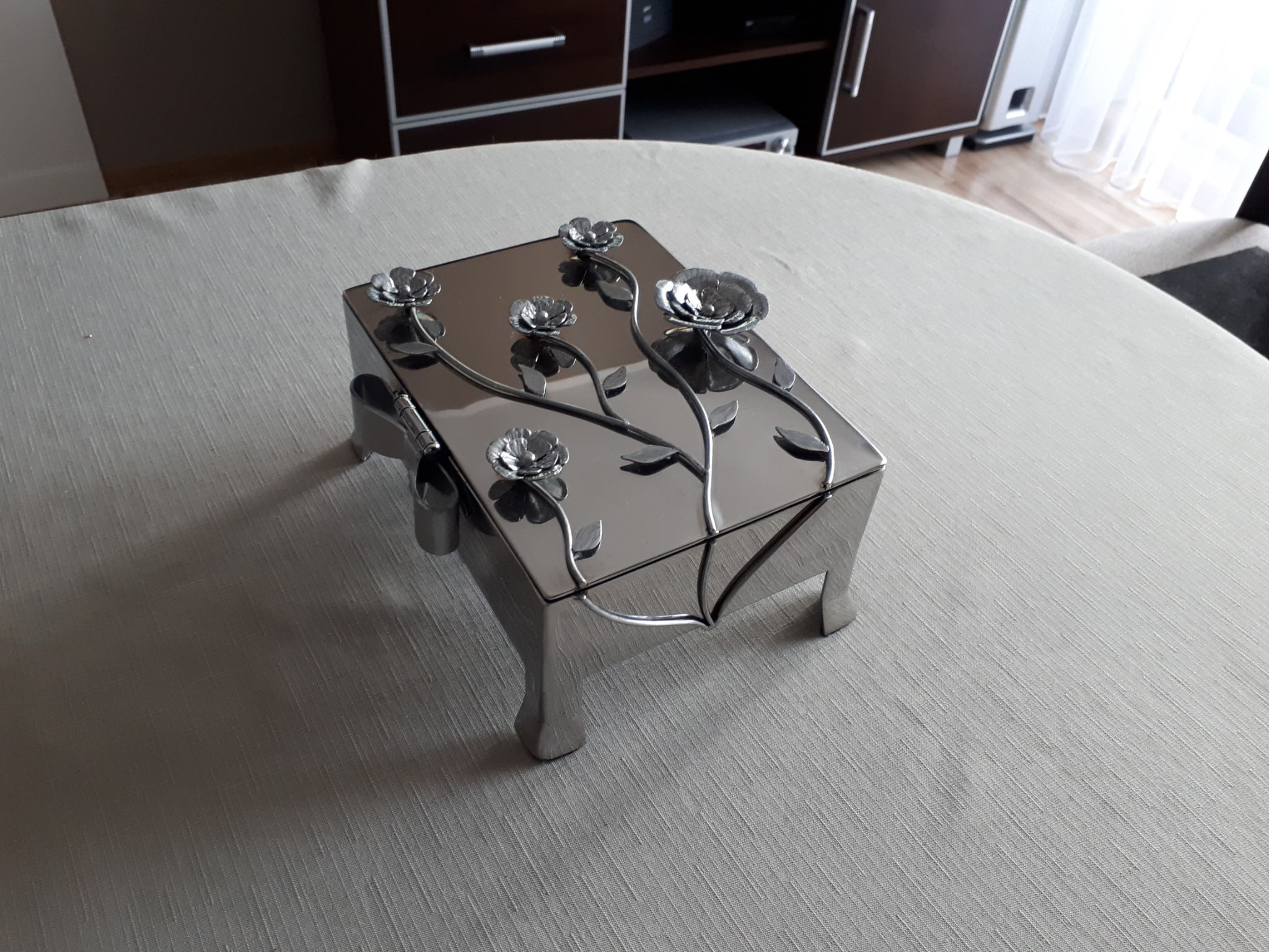 Dekoracyjna, metalowa szkatułka z motywem kwiatowym na blacie stołu, odbijająca światło. Widoczne elementy wystroju wnętrza w tle.