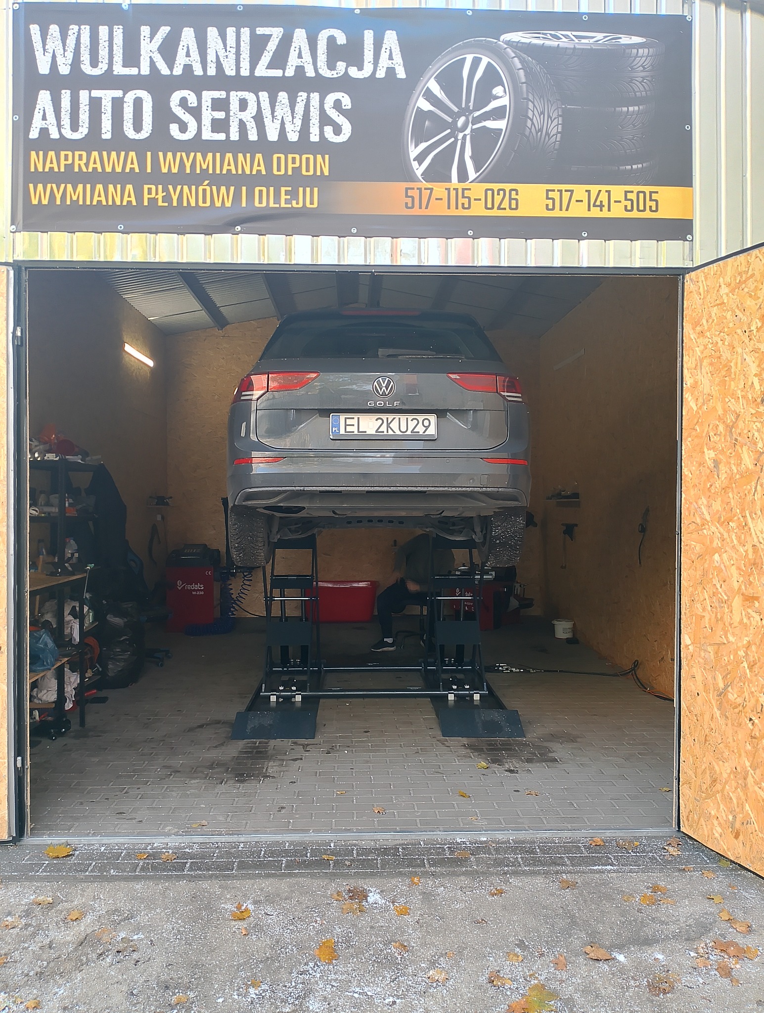 Szary Volkswagen Golf na podnośniku w warsztacie wulkanizacyjnym z otwartymi drzwiami. Widoczny baner reklamowy z numerami telefonów.
