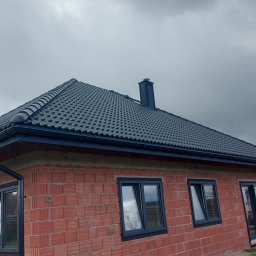 DACHY Krystian Witecki - Dom z nowym, grafitowym dachem z dachówki ceramicznej i rynnami w kolorze. Widoczny komin i fragment elewacji z cegły. Pochmurne niebo w tle.