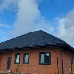Nowoczesny dom z czerwoną cegłą i czarnym dachem z dachówki, ujęcie z dołu na tle błękitnego nieba z chmurami. Widoczne czarne rynny i okna.