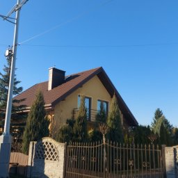 DACHY Krystian Witecki - Dom jednorodzinny z brązowym dachem i kominem, ogrodzony metalowym płotem. Widoczne drzewa iglaste i latarnia uliczna. Błękitne niebo w tle.
