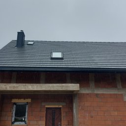 DACHY Krystian Witecki - Ciemnoszary dach z dachówki bitumicznej z oknami połaciowymi i czarną rynną na surowym budynku z czerwonej cegły. Widok z dołu, pochmurne niebo.