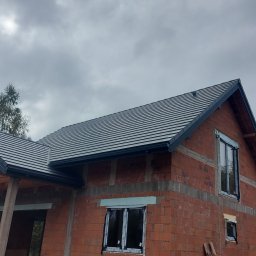 DACHY Krystian Witecki - Surowy budynek z ciemnoszarą dachówką karpiówką i czarnymi rynnami, widok z dołu na tle pochmurnego nieba i drzew. Częściowo widoczna osoba w czerwonej czapce.