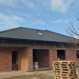 DACHY Krystian Witecki - Dom w trakcie budowy z widocznym dachem z dachówką ceramiczną i oknem dachowym. Ściany z czerwonej cegły, palety budowlane na pierwszym planie. Błękitne niebo w tle.