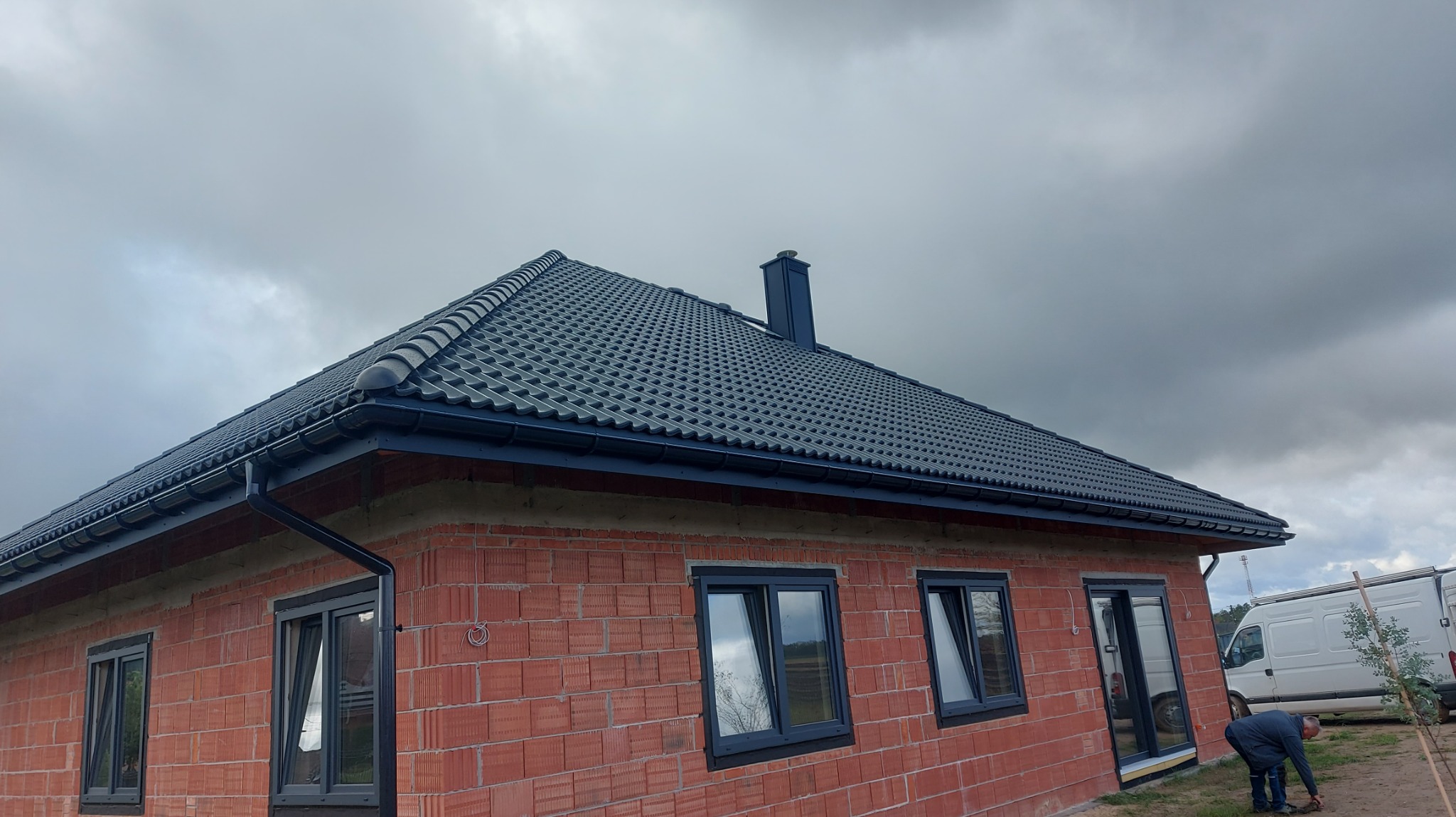 Dom z nowym, grafitowym dachem z dachówki ceramicznej i rynnami w kolorze. Widoczny komin i fragment elewacji z cegły. Pochmurne niebo w tle.