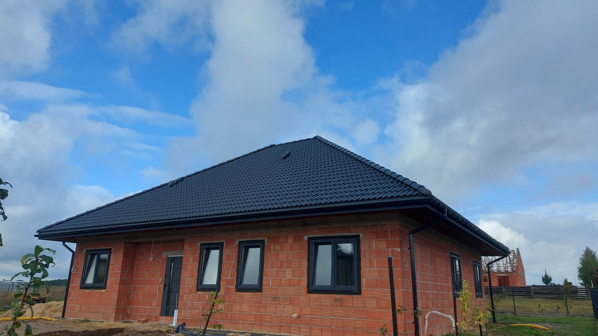 Nowoczesny dom z czerwoną cegłą i czarnym dachem z dachówki, ujęcie z dołu na tle błękitnego nieba z chmurami. Widoczne czarne rynny i okna.