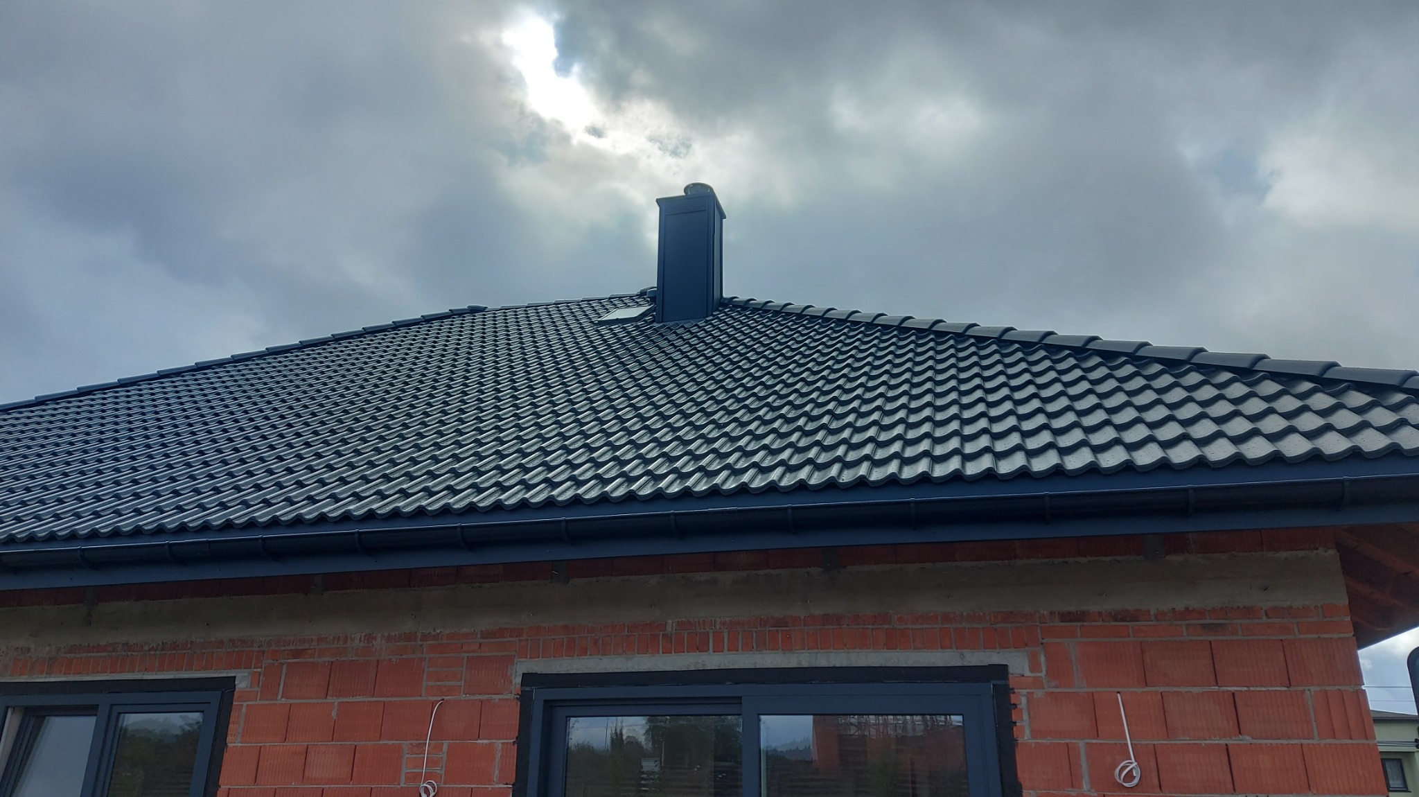 Ciemny dach z dachówki ceramicznej na nowym domu w trakcie budowy, z widocznym kominem i oknami. Pochmurne niebo w tle.