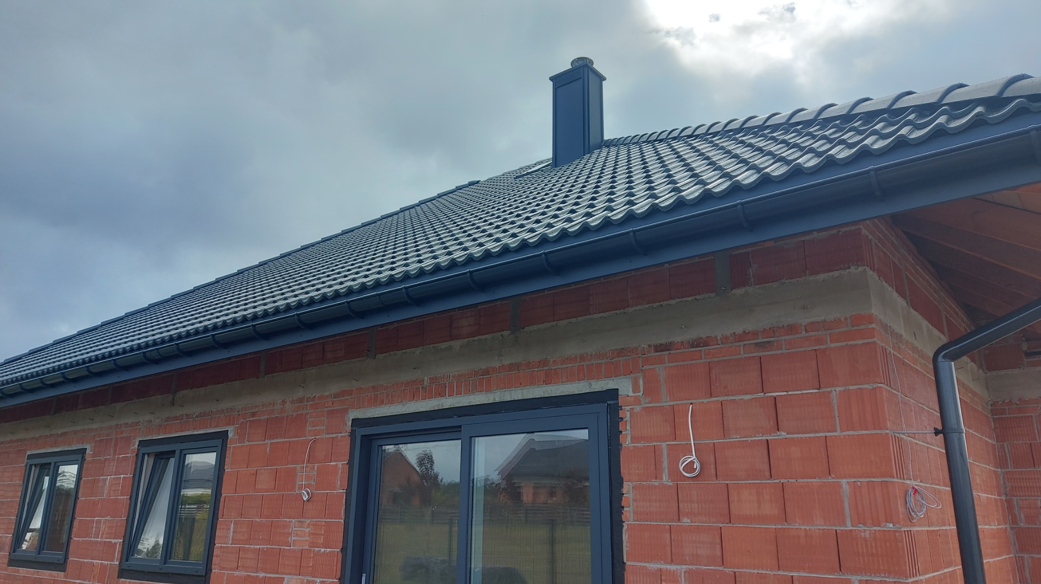 Ciemna dachówka ceramiczna na nowym domu z cegły. Widoczne rynny w kolorze dachu i komin. Fragment elewacji z oknami i wystającymi kablami.
