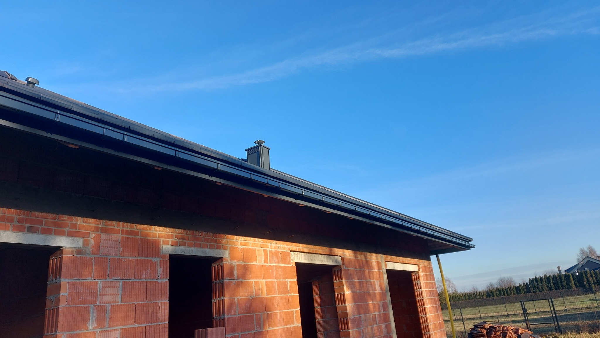 Ceglany budynek w trakcie budowy z zamontowanymi czarnymi rynnami i dachem, widok z dołu na tle błękitnego nieba.
