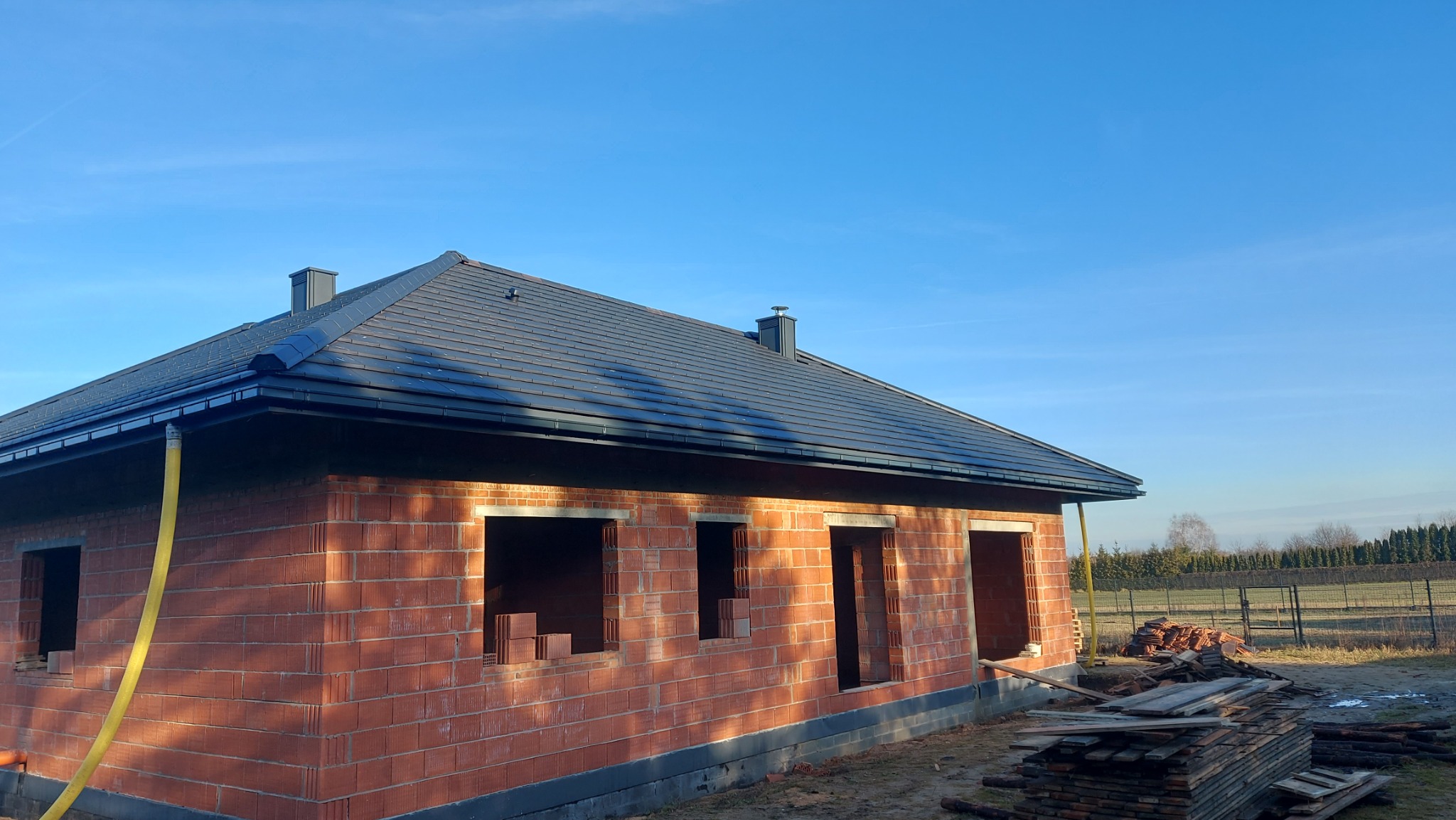 Surowy budynek z ciemną dachówką i rynnami w słoneczny dzień. Widoczne cegły, okna bez szyb i ułożone deski. Dach pokryty, kominy zamontowane.
