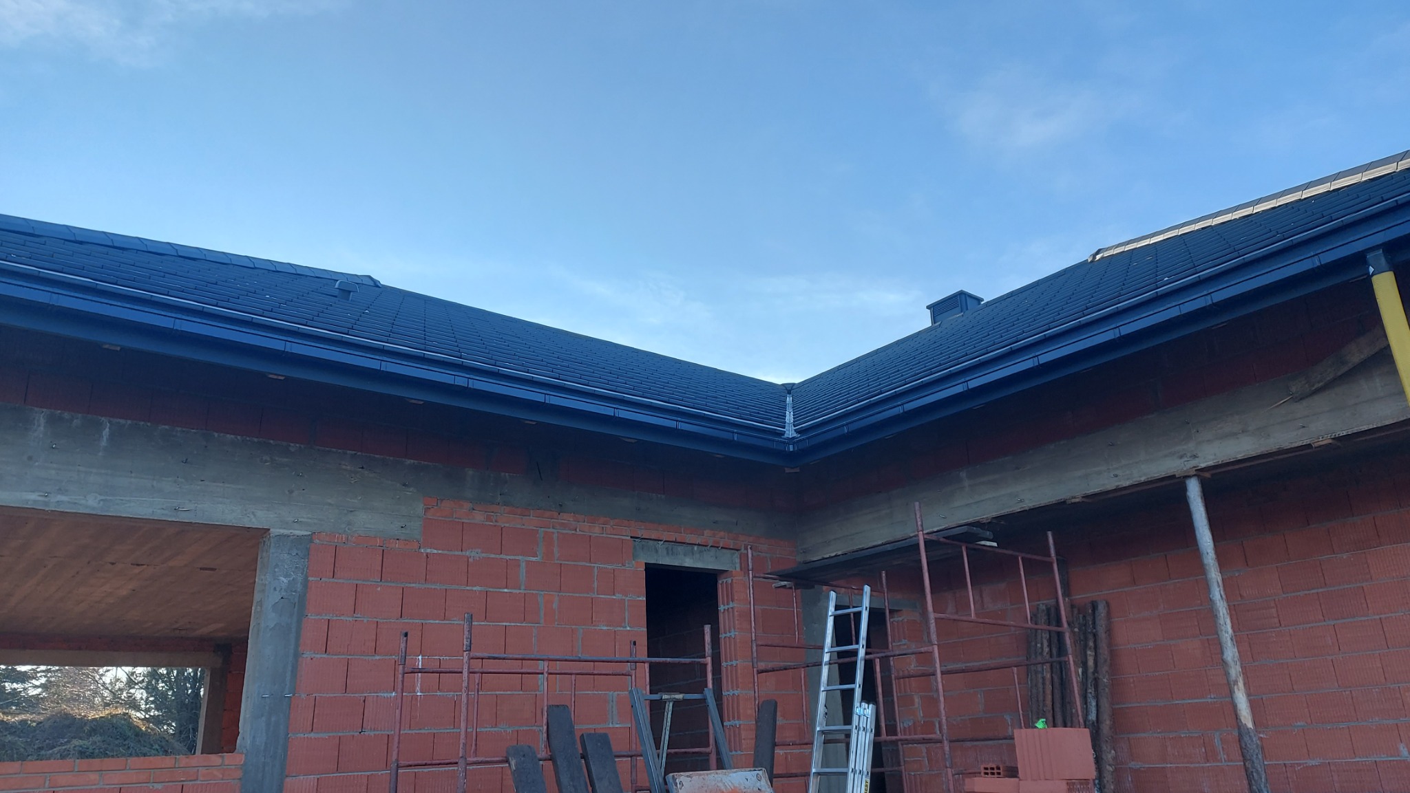 Ciemny dach z rynnami na surowym budynku z czerwonej cegły. Widoczne rusztowania i drabina. Błękitne niebo w tle. Ujęcie z dołu.