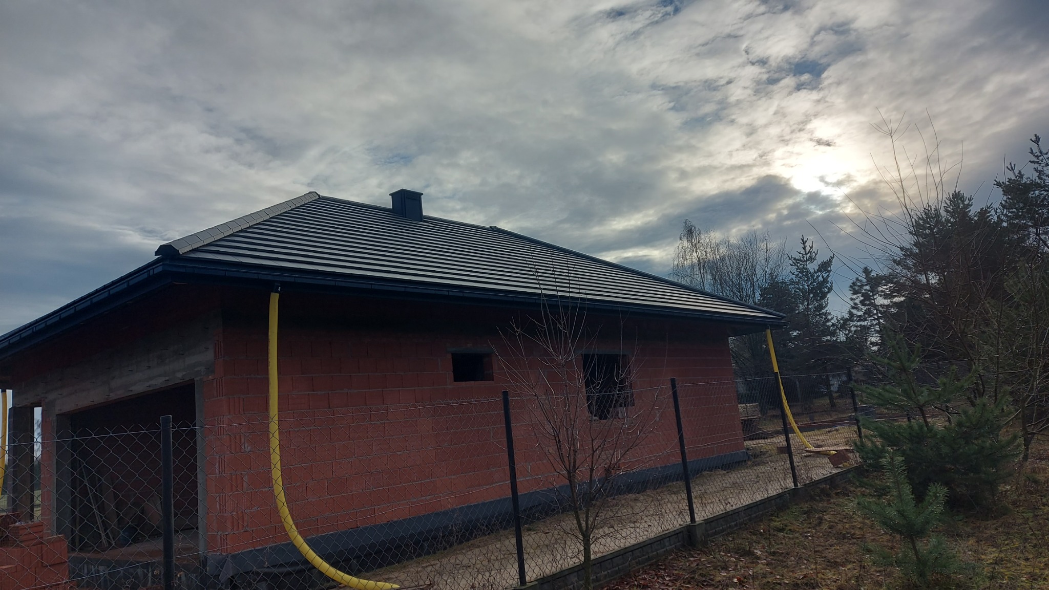 Dom w trakcie budowy z czarnym dachem i rynnami. Widoczne cegły, ogrodzenie i żółty wąż. Pochmurne niebo w tle.