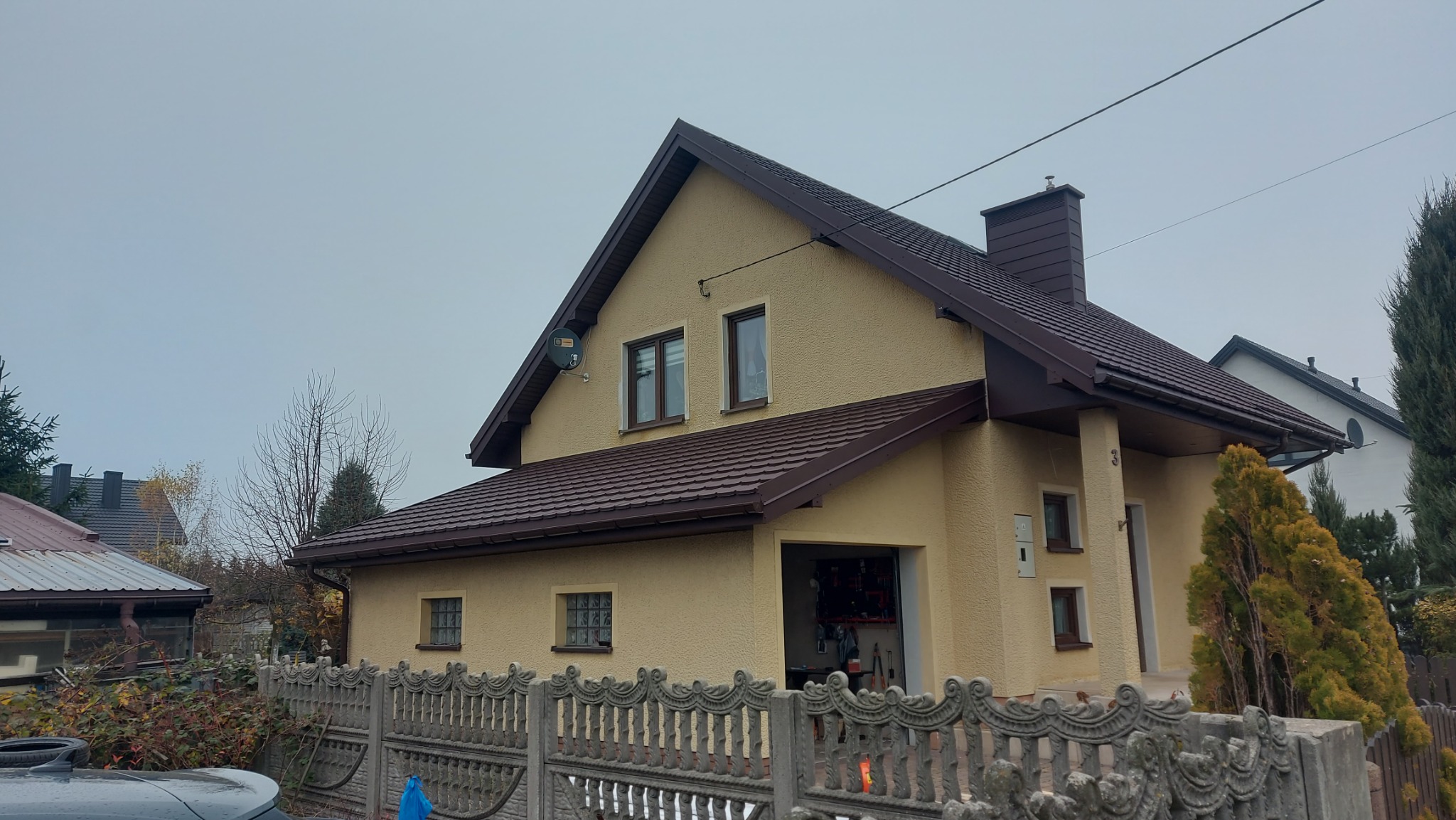Dom jednorodzinny z brązowym dachem i rynnami, widoczny zza ozdobnego, betonowego ogrodzenia. Elewacja w odcieniach beżu, garaż otwarty, pochmurne niebo w tle.