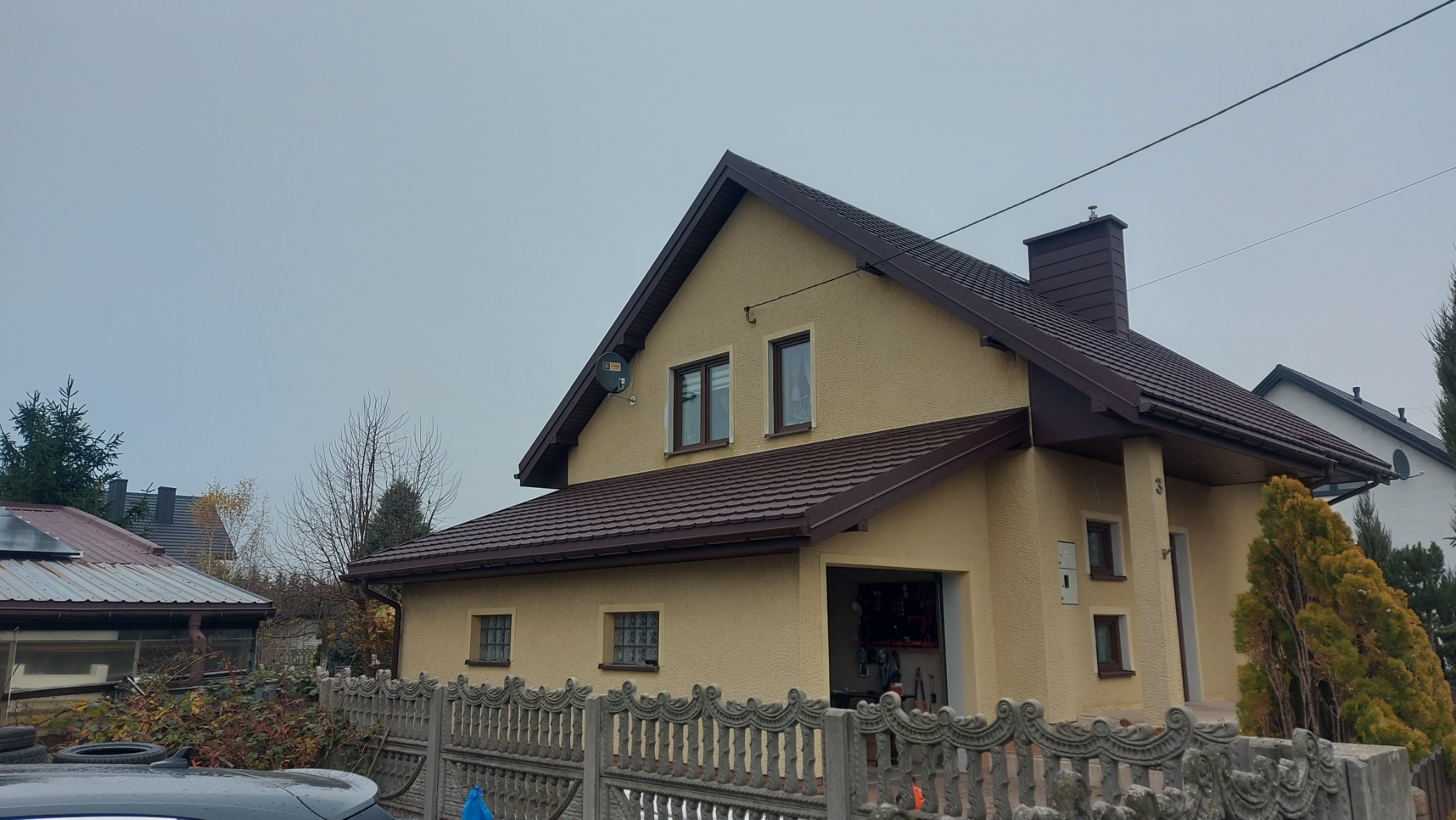 Dom jednorodzinny z brązowym dachem i rynnami w Łasku. Widoczny fragment ogrodzenia i garaż. Ujęcie z dołu, pochmurne niebo w tle.