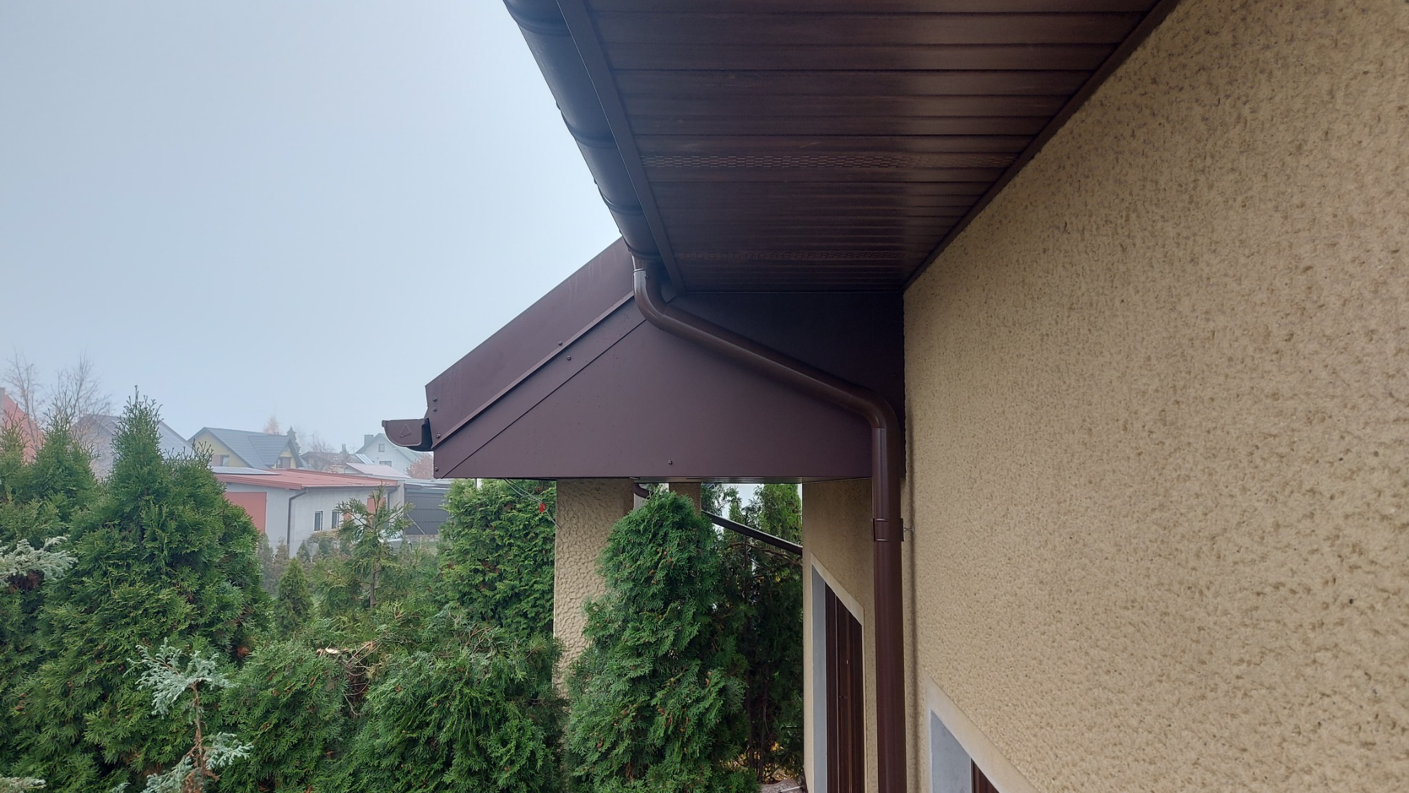 Brązowa rynna spustowa i okap dachu na tle beżowej elewacji domu, widok z dołu, zieleń w tle, pochmurny dzień.