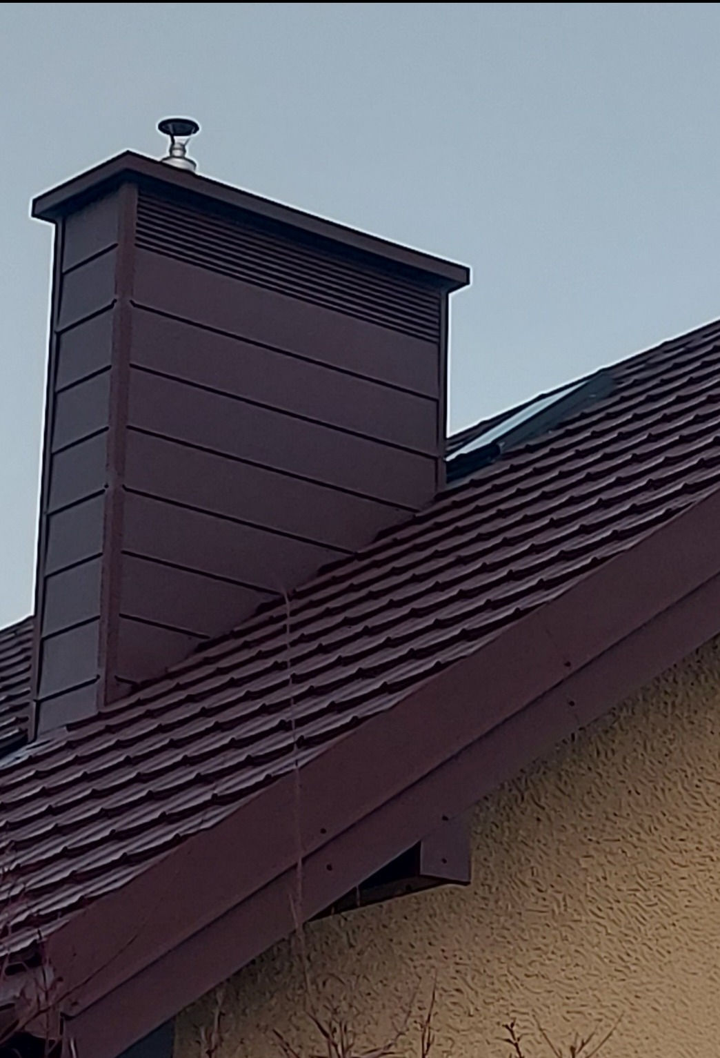 Bordowy komin wentylacyjny z metalową czapką na dachu z bordowej blachodachówki, widok z dołu na tle jasnego nieba.