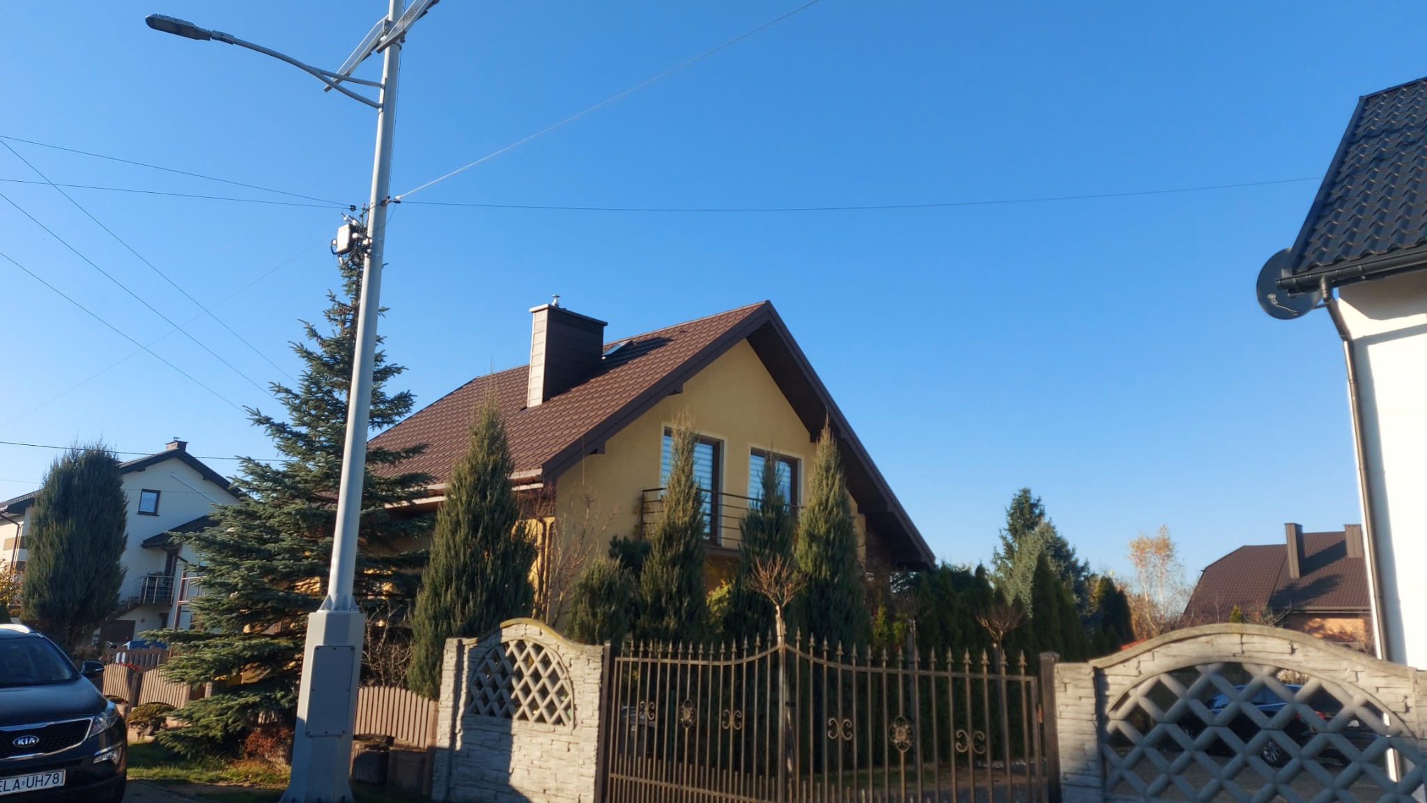 Dom jednorodzinny z brązowym dachem i kominem, ogrodzony metalowym płotem. Widoczne drzewa iglaste i latarnia uliczna. Błękitne niebo w tle.