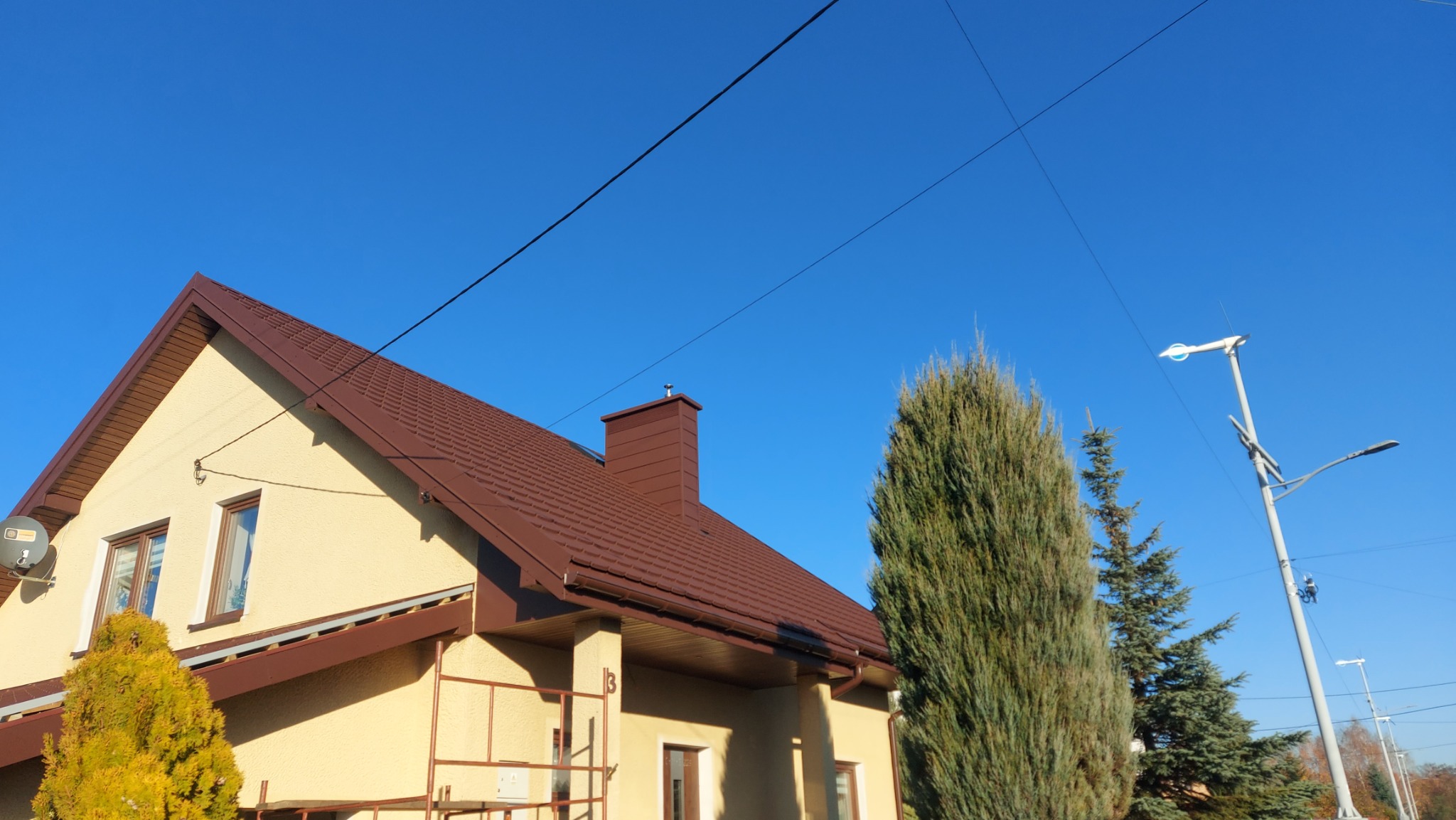Dom jednorodzinny z brązowym dachem i kominem, sfotografowany w słoneczny dzień na tle błękitnego nieba. Widoczne drzewa iglaste i fragment rusztowania.