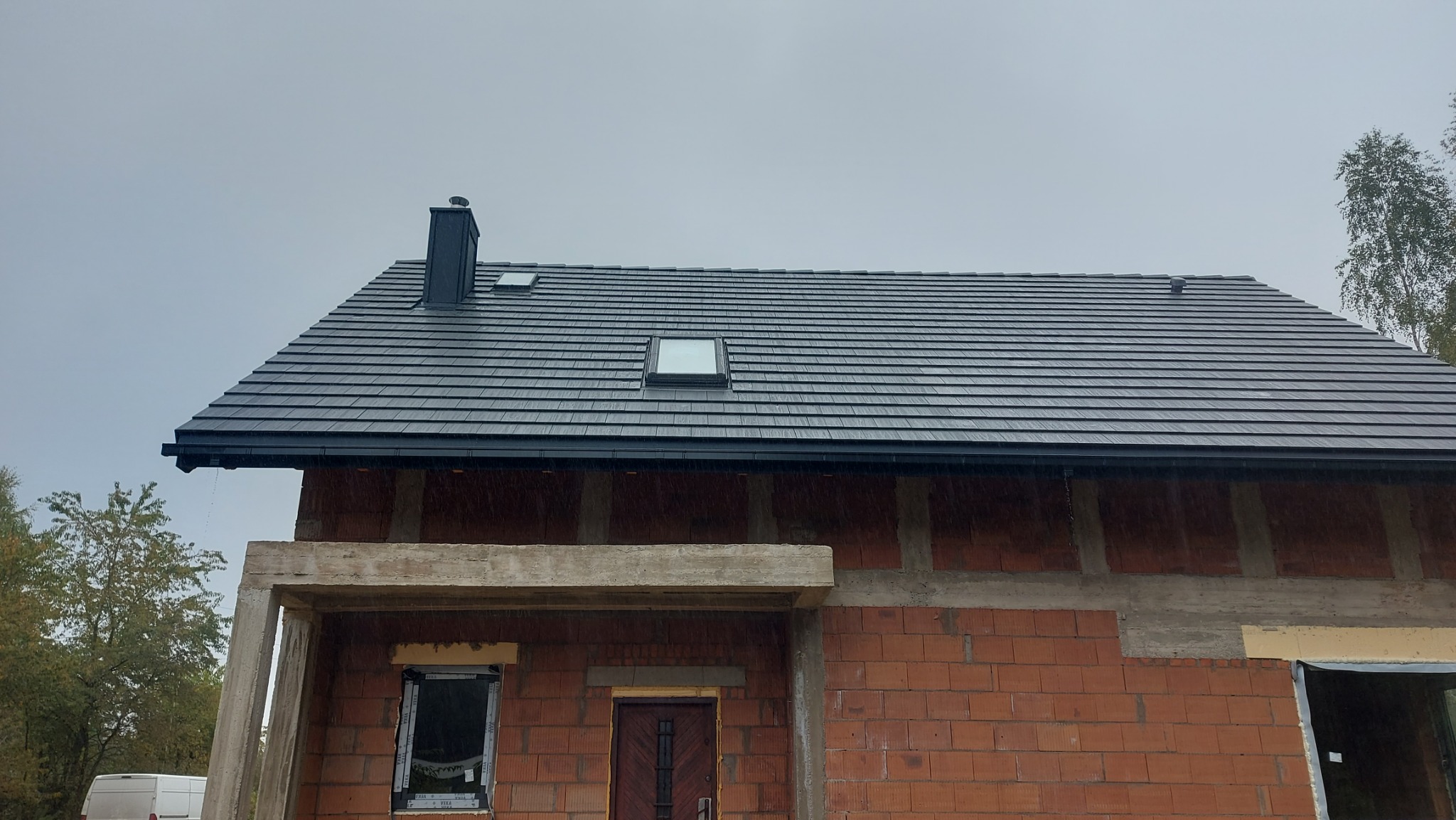 Ciemnoszary dach z dachówki bitumicznej z oknami połaciowymi i czarną rynną na surowym budynku z czerwonej cegły. Widok z dołu, pochmurne niebo.