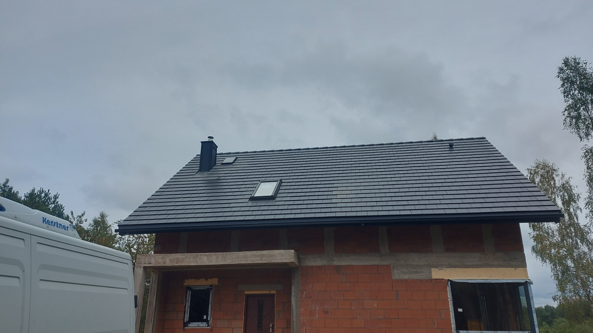 Nowy, grafitowy dach z dachówką ceramiczną i oknami połaciowymi na surowym budynku z czerwonej cegły. Widoczny fragment białego samochodu dostawczego.