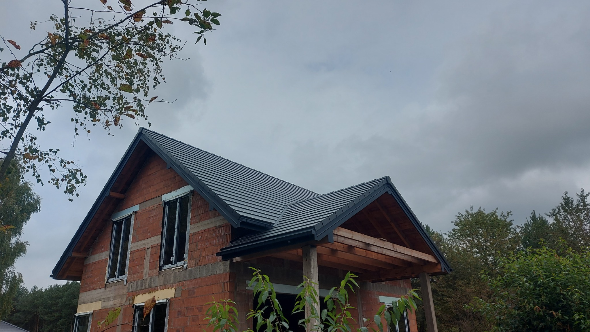 Dom w trakcie budowy z nowym, grafitowym dachem z dachówki ceramicznej. Widoczne rynny i obróbki blacharskie. Częściowo ocieplone okna. Pochmurne niebo w tle.